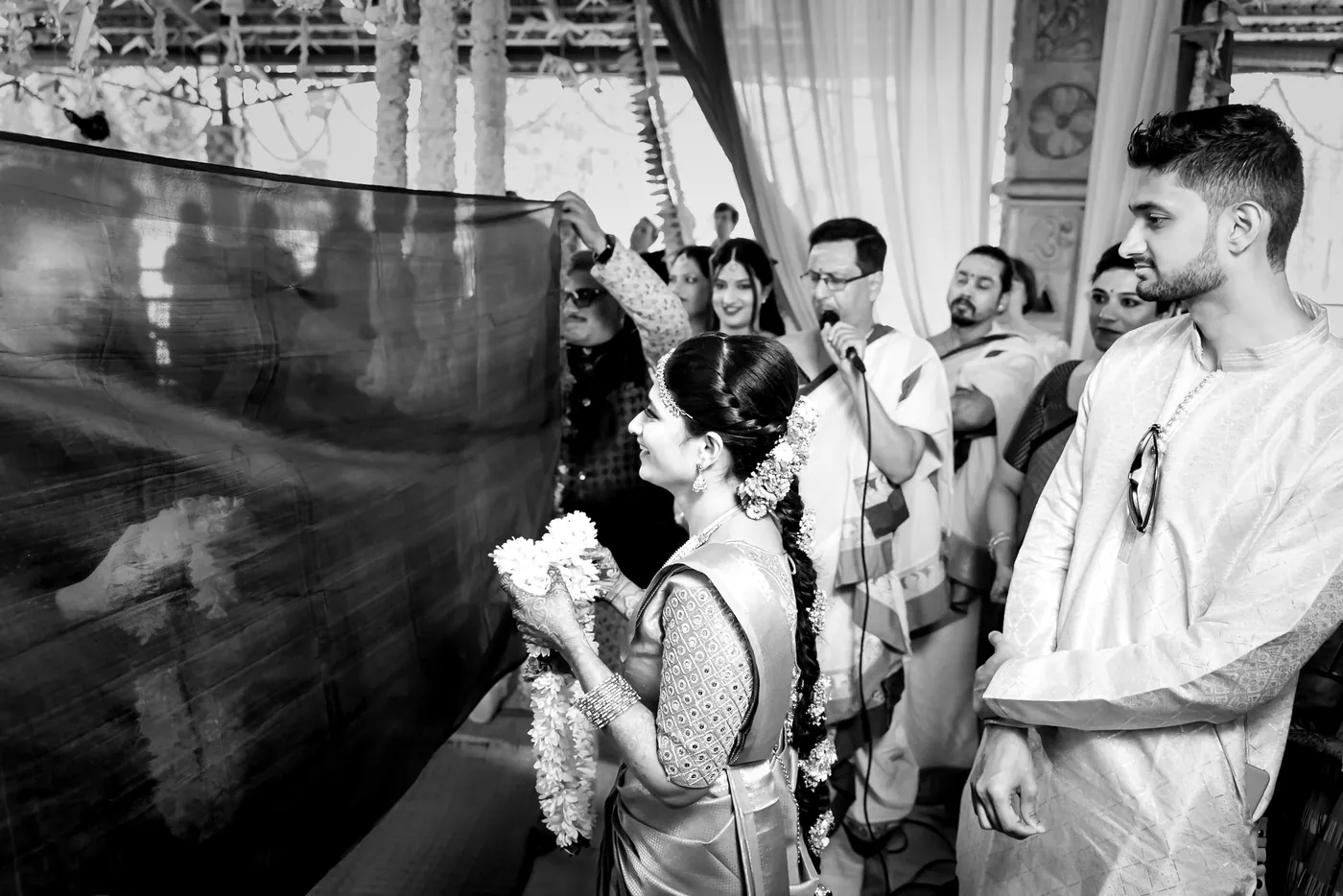 Medha holds a jasmine garland behind a raised cloth screen while a priest speaks into a microphone