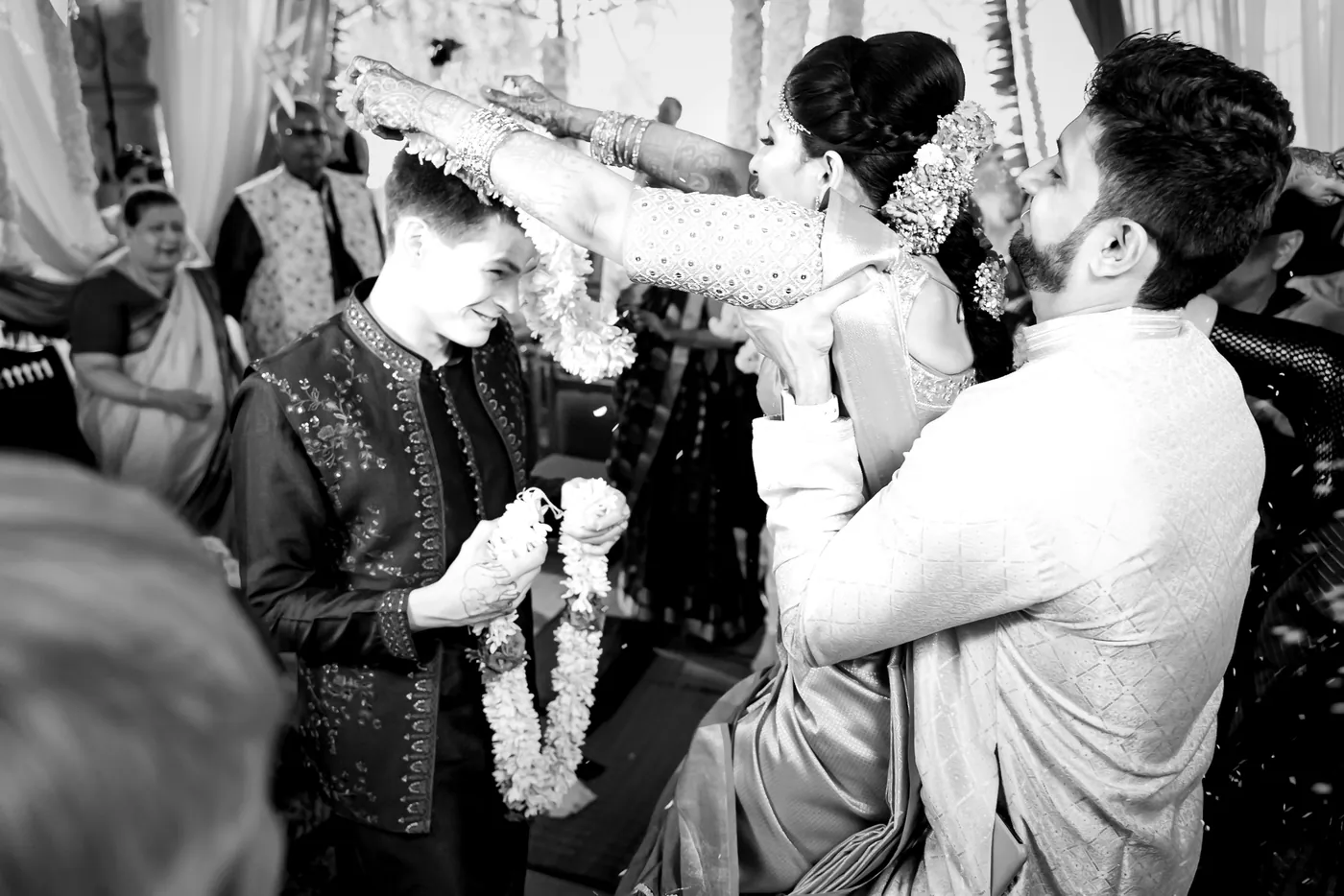 A man in cream lifts Medha toward Stefan, who laughs while holding his own jasmine garland