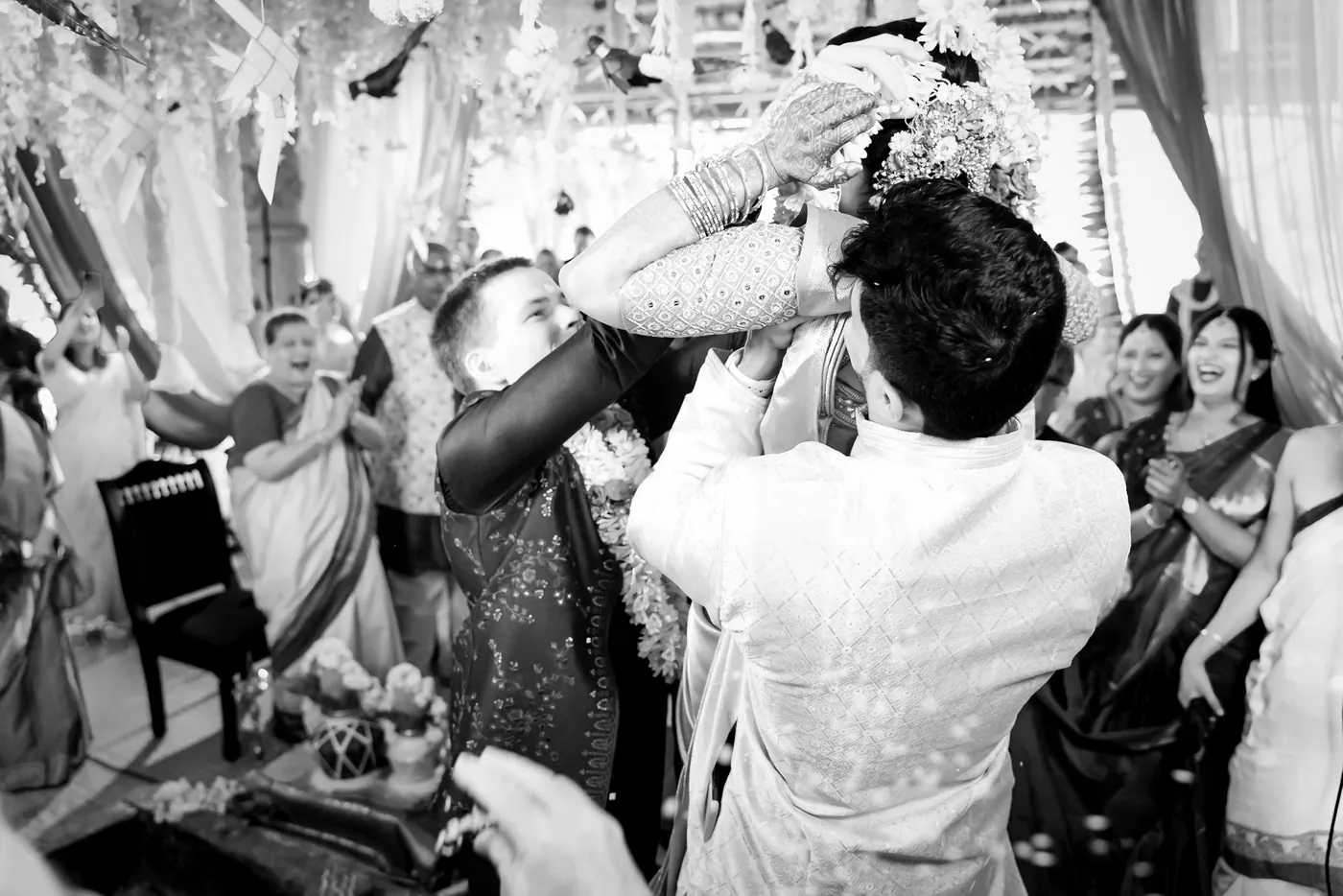 A man in cream lifts Medha high to garland Stefan while guests laugh and clap around them