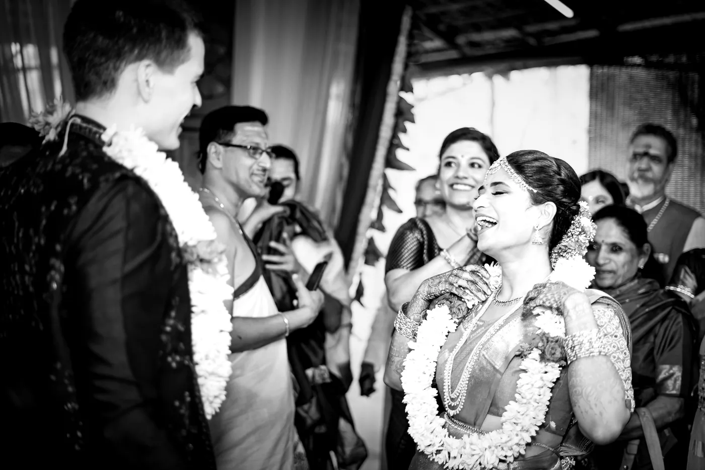 Black and white of Stefan and Medha in jasmine garlands laughing as guests and a man with glasses look on
