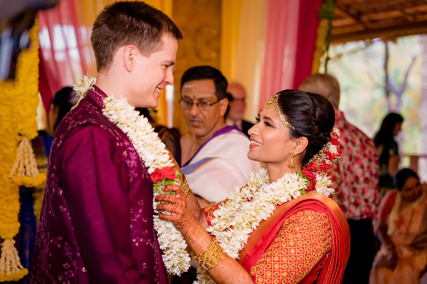 Medha and Stefan smile at each other wearing jasmine garlands under yellow and pink drapes