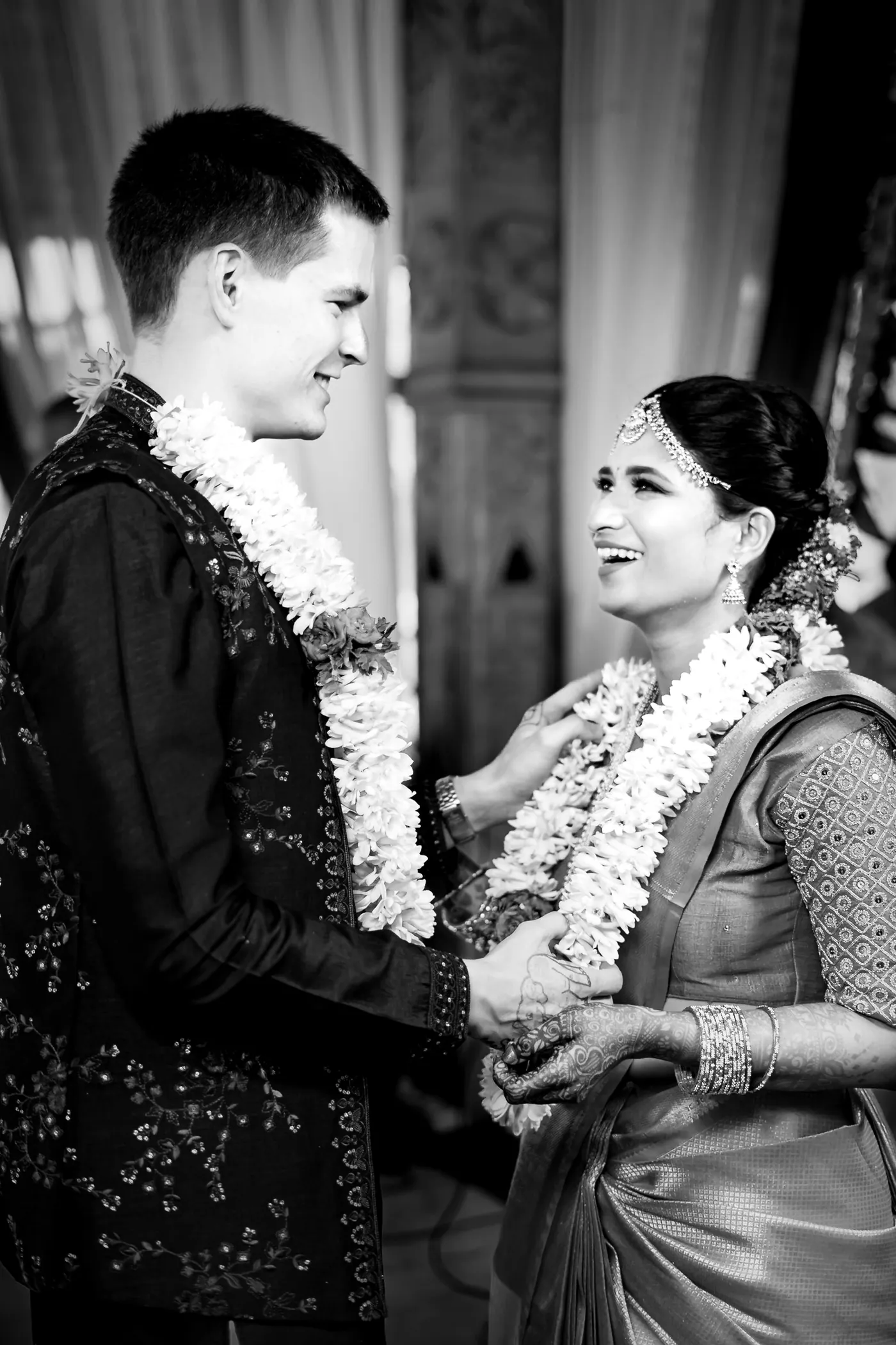 Black and white of Stefan smiling down at Medha, both in garlands, as she laughs and holds his hand