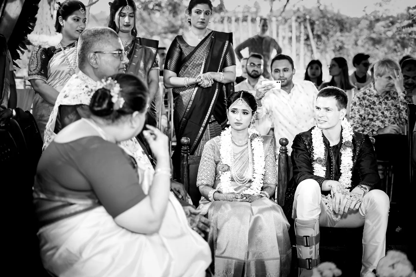 A woman in the foreground touches her face, seemingly in tears, as garlanded Medha and Stefan sit behind