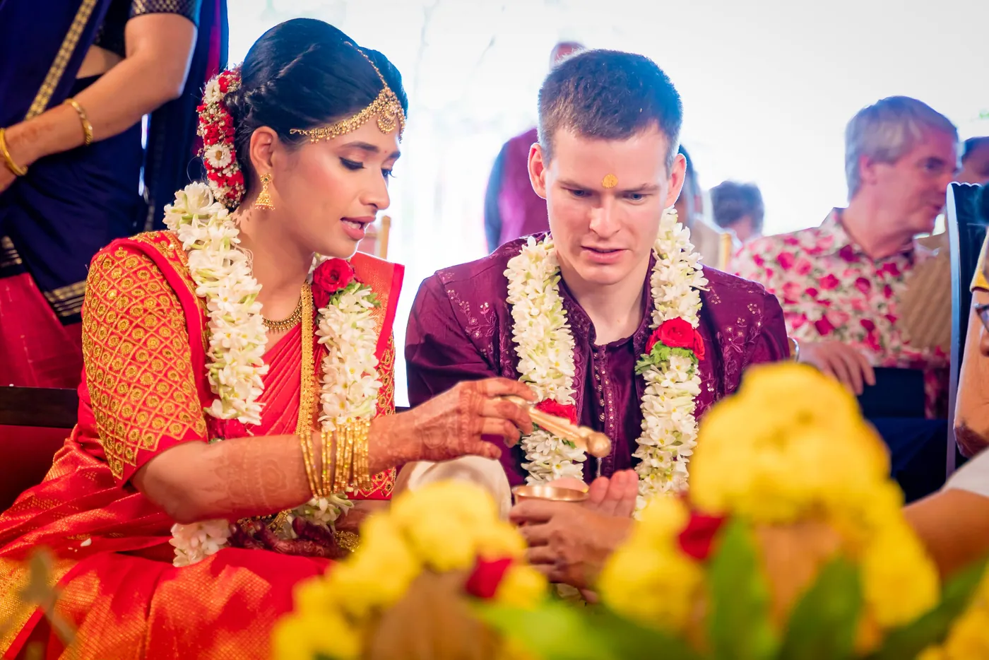 Medha and Stefan in jasmine garlands look down at their hands over bright yellow and red flowers