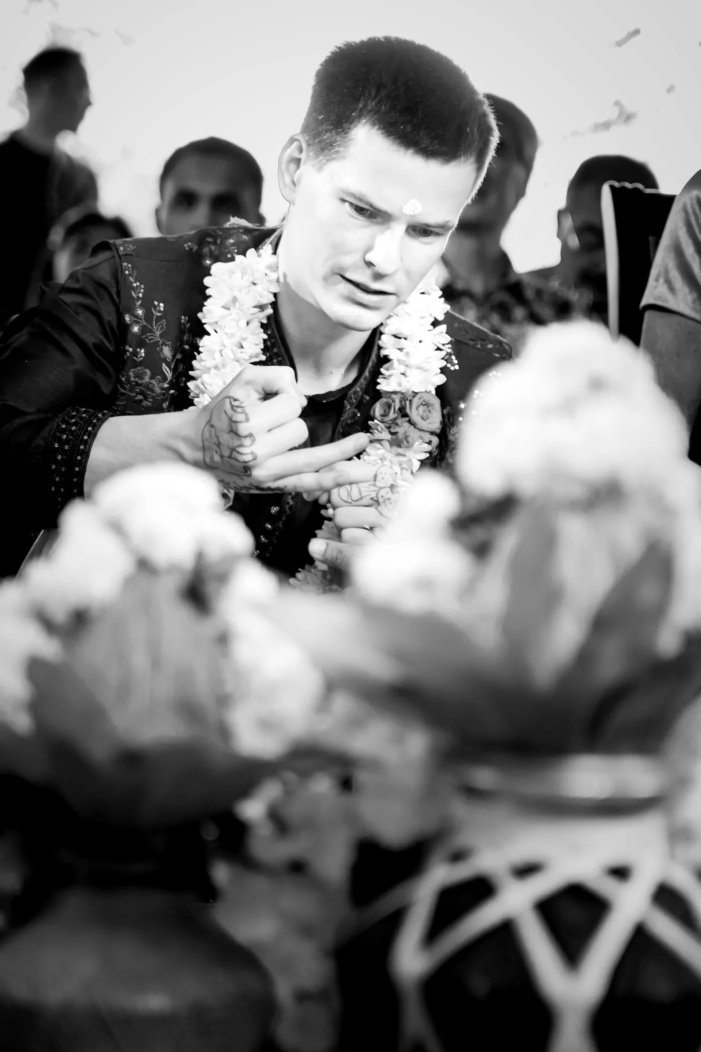 Black and white of Stefan in a jasmine garland looking down at his hands among blurred flowers and clay pots