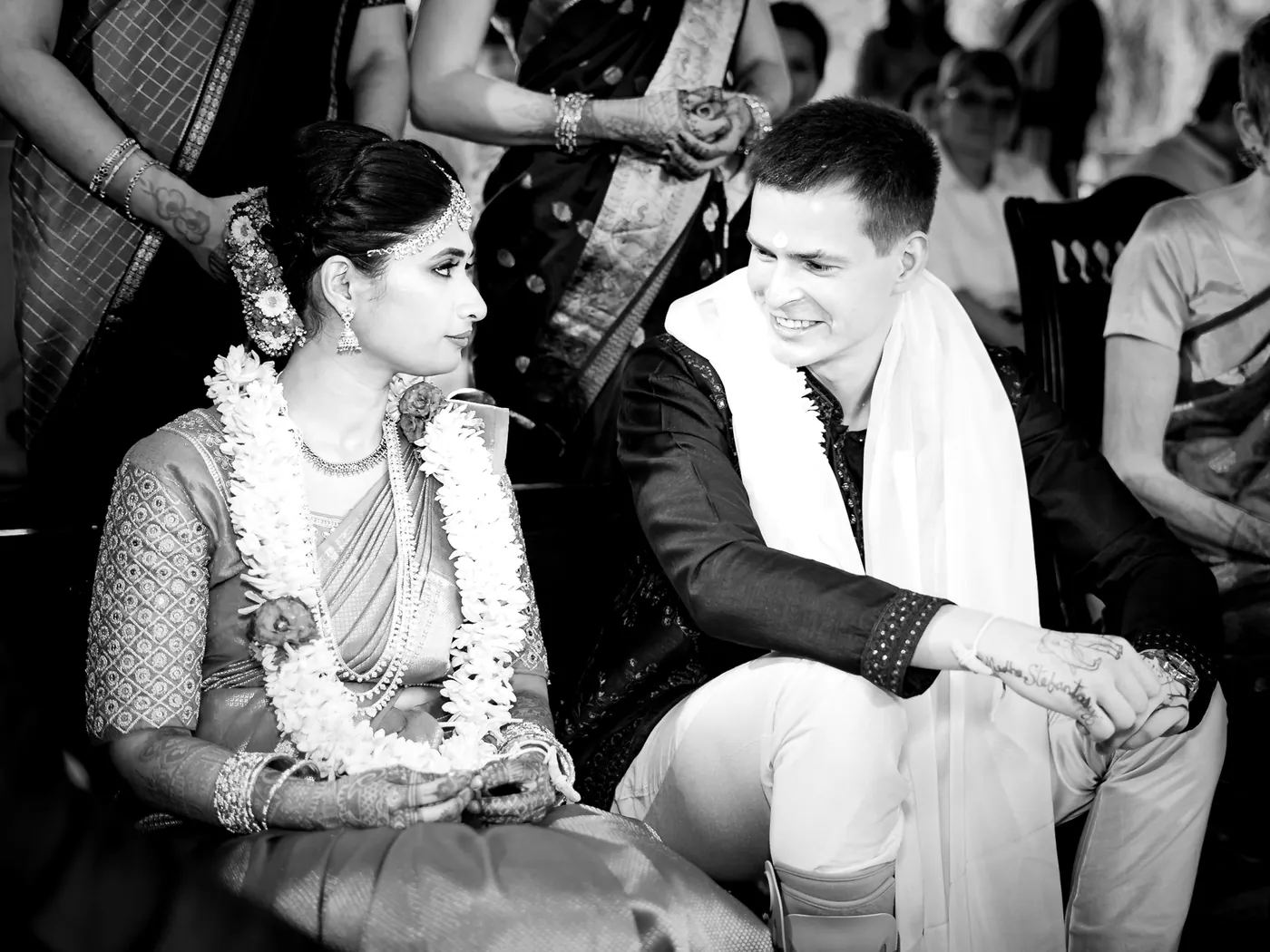 Black and white of garlanded Medha and Stefan seated close, glancing at each other with a smile