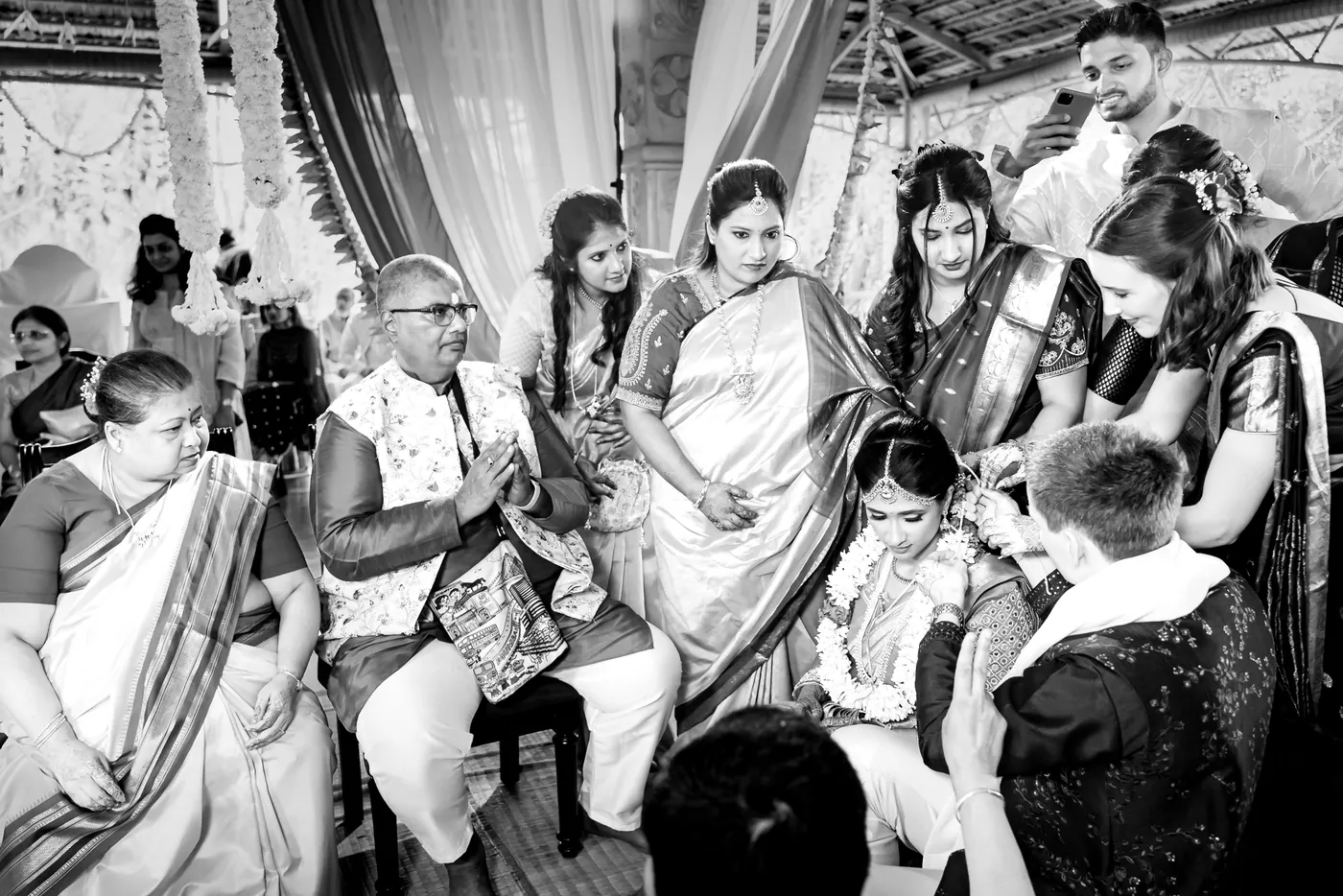 Family crowd close around the garlanded couple as a man presses his palms together and others lean in