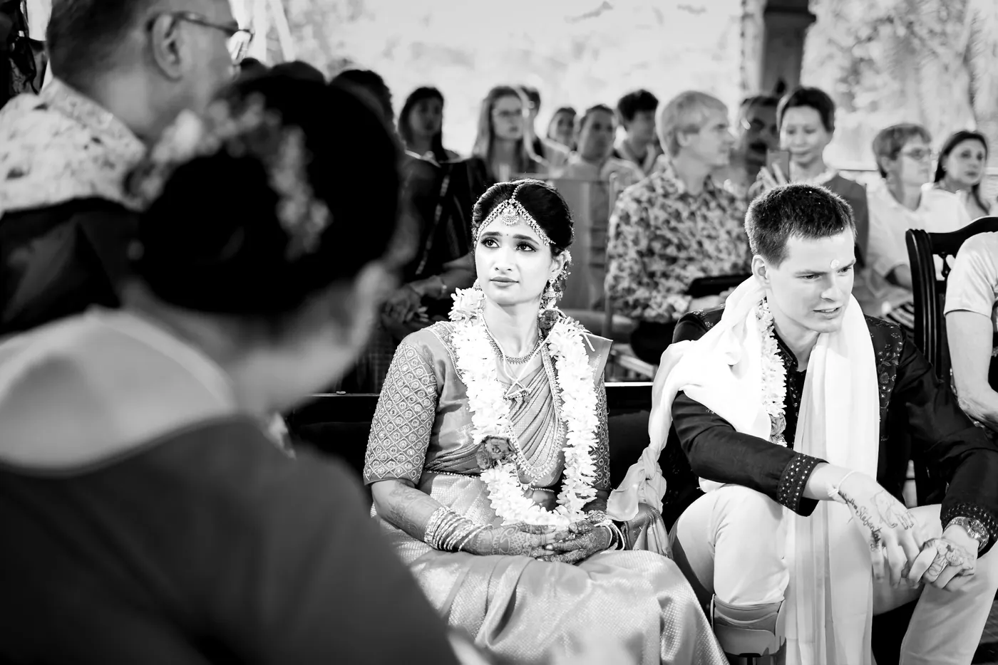 Black and white of Medha and Stefan seated side by side in garlands as a guest leans in