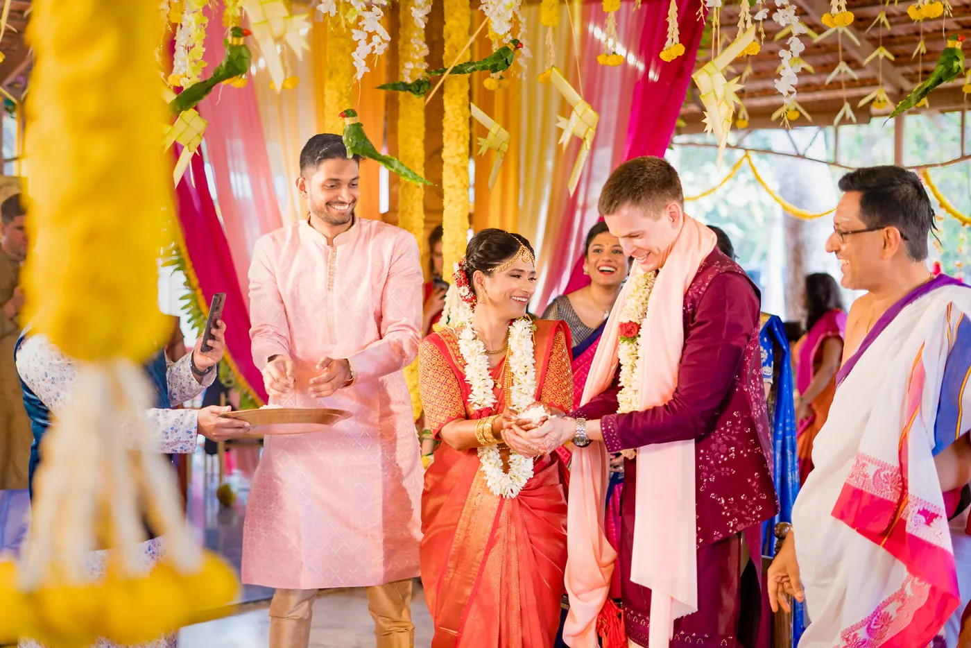 Medha and Stefan in garlands join hands with a priest and two men standing under yellow and pink drapes