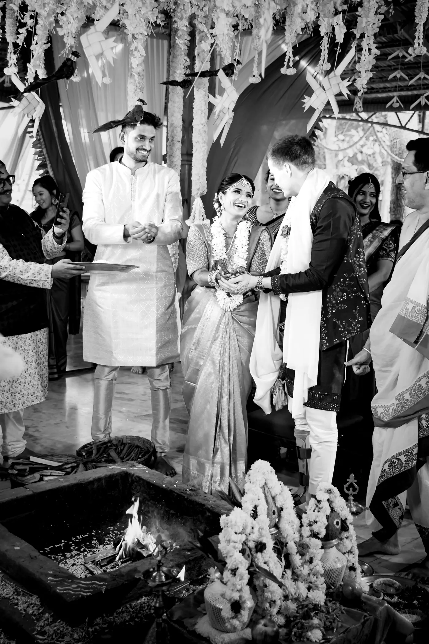 Black and white of Medha and Stefan standing by a small fire as a priest and a man watch