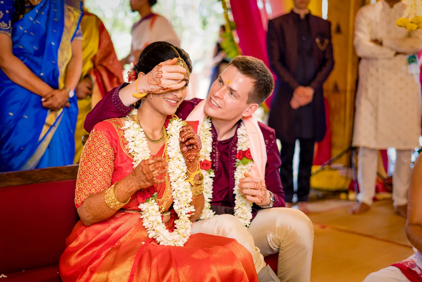 A hand covers Medha's eyes as Stefan leans in smiling, both in jasmine garlands, guests standing behind