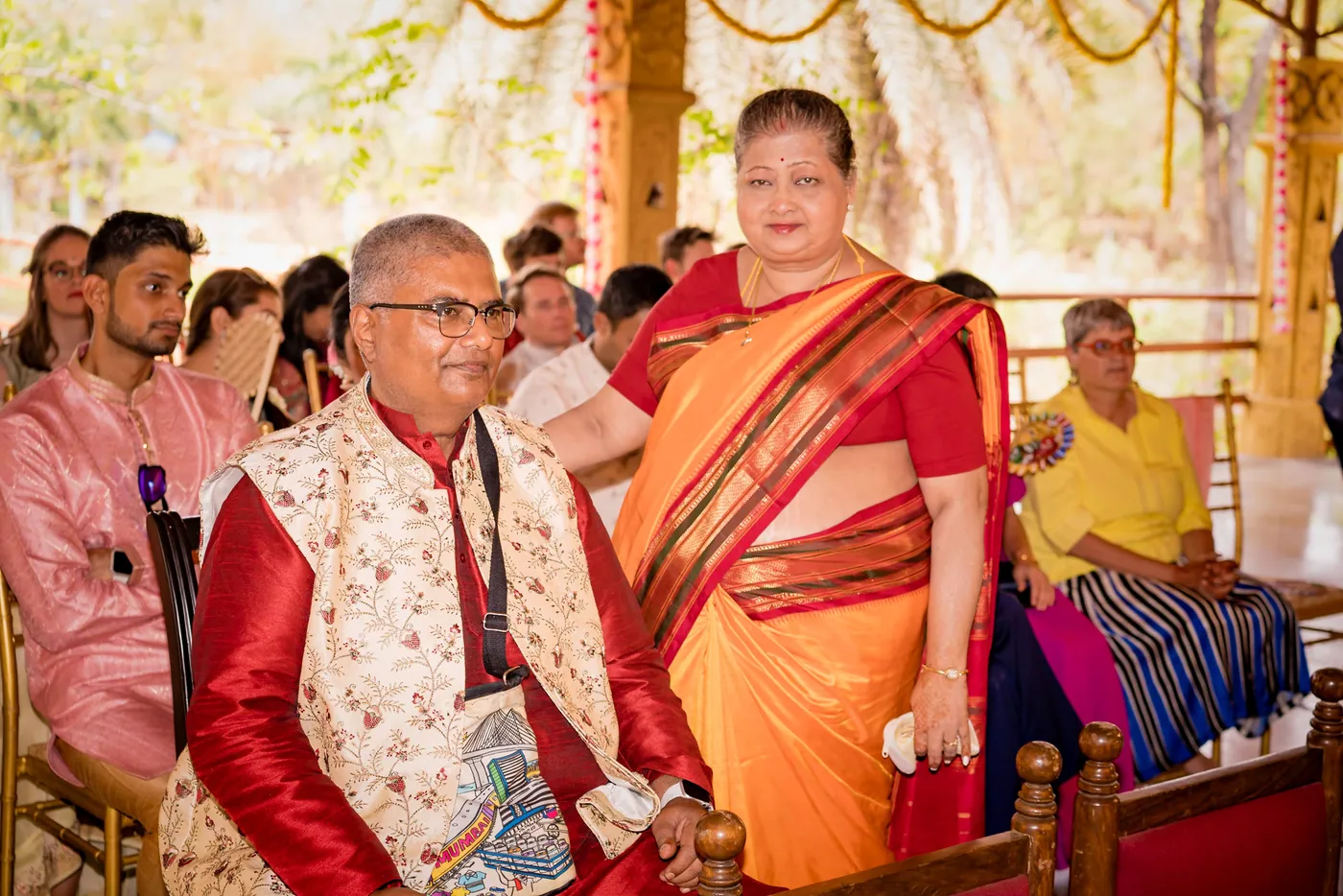 A man in a red and white waistcoat and a woman in an orange sari sit in the audience