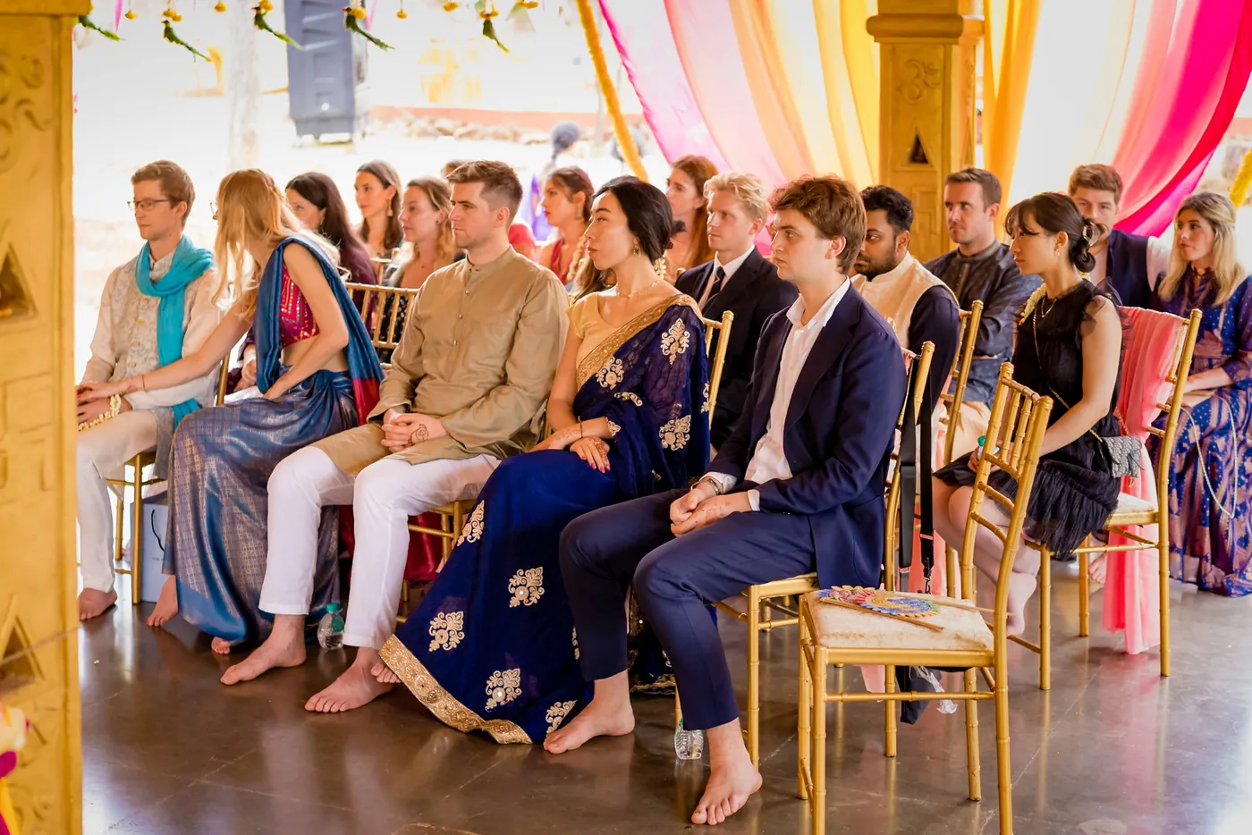 Rows of guests in dark and bright clothes sit barefoot on a polished floor