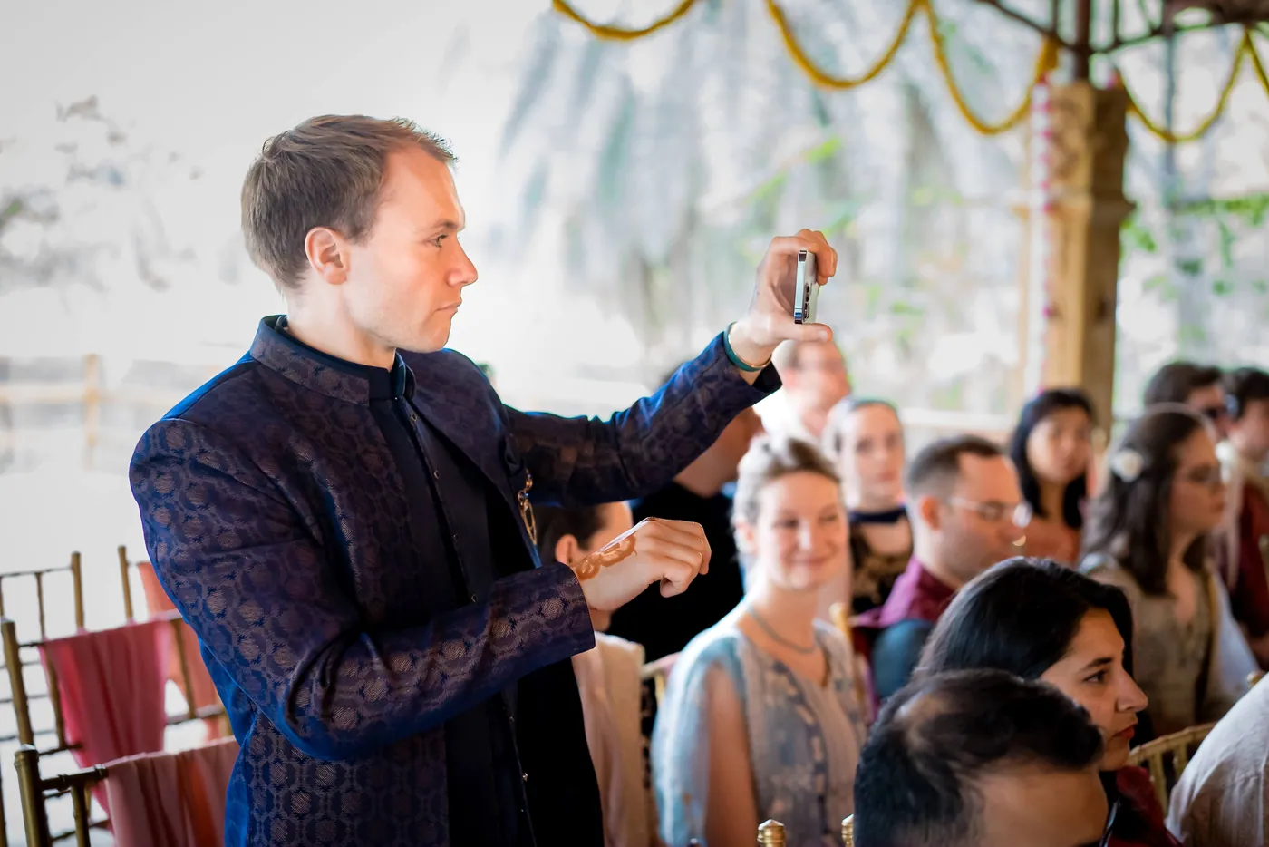 A man in a navy kurta takes a photo with his phone as guests sit behind him