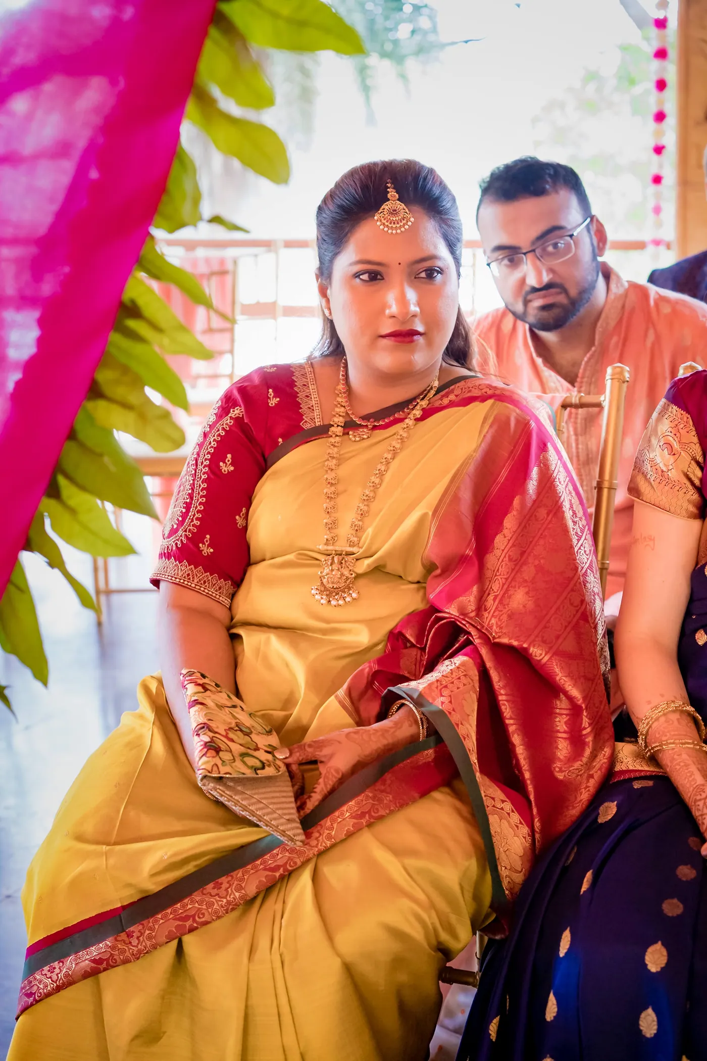 A woman in a yellow sari with a red border sits beside a man in an orange kurta