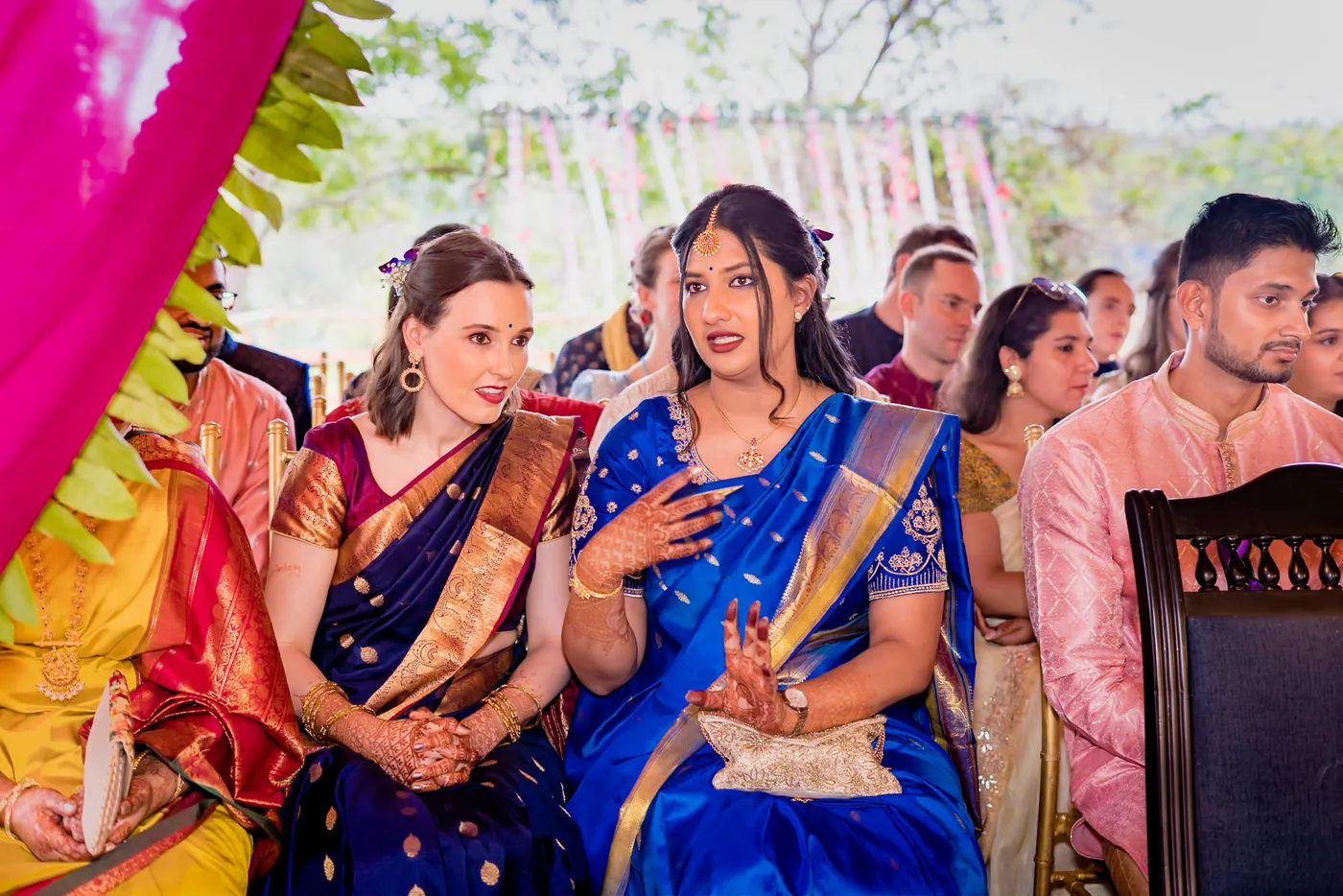Two women in blue saris whisper to each other among the audience