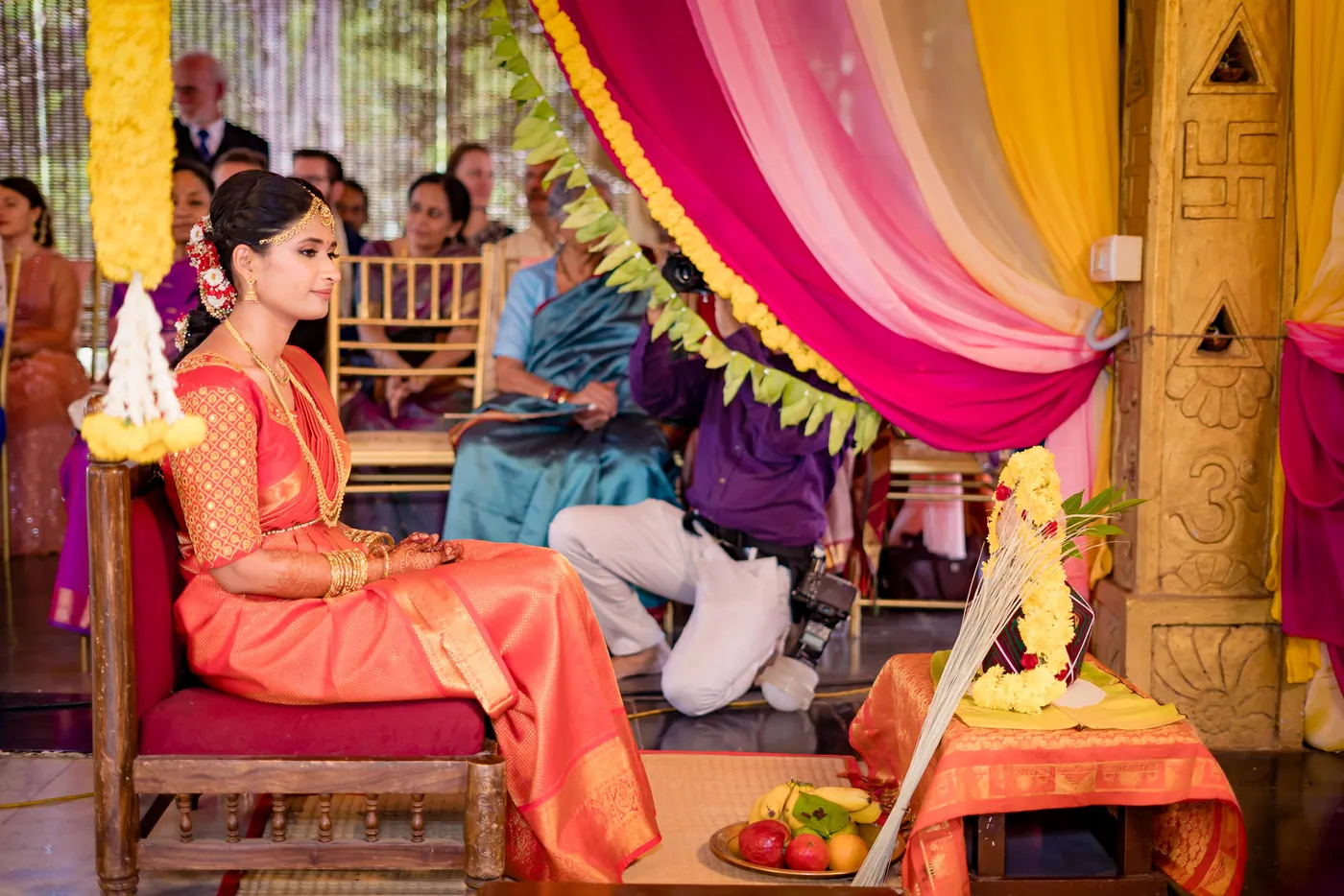 Medha sits in an orange sari on a carved chair beside decorated ceremonial pots