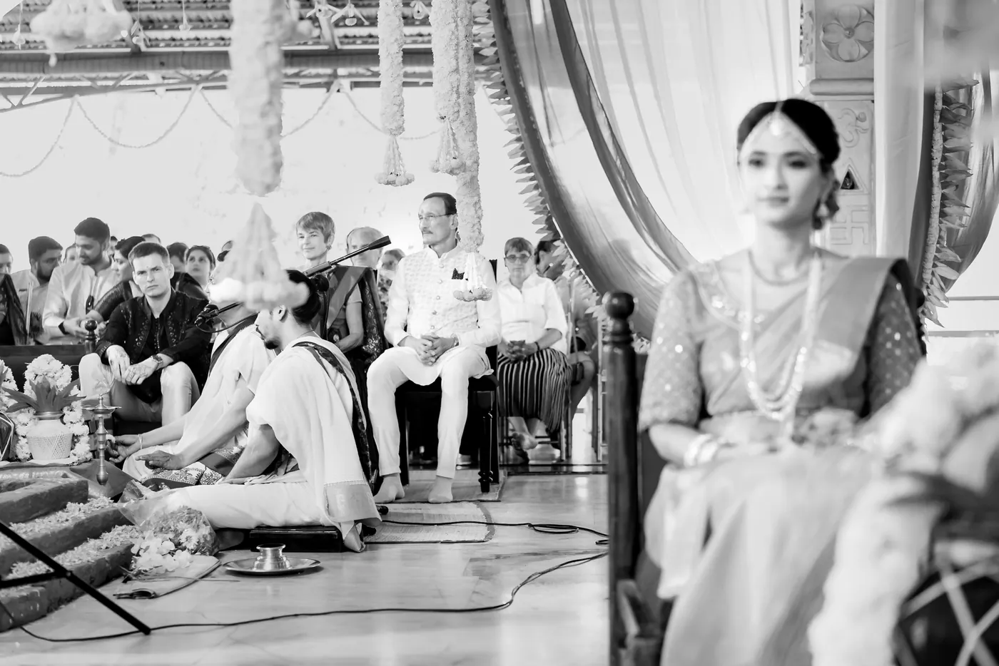 Black and white of Medha seated at the ceremony as guests sit on the floor behind her