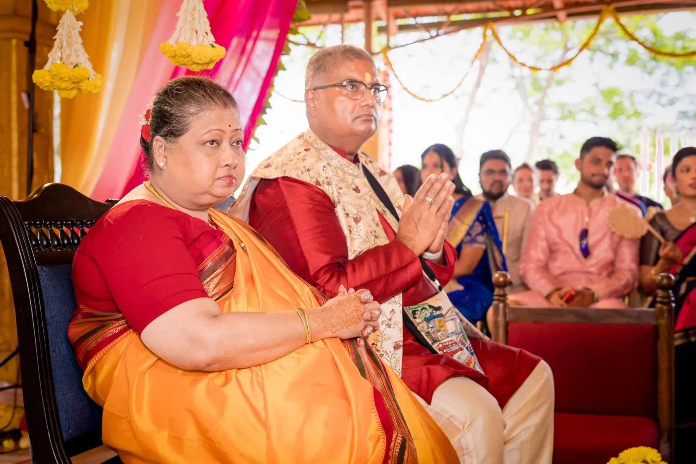 A woman in orange and a man in a gold waistcoat sit among guests at the ceremony
