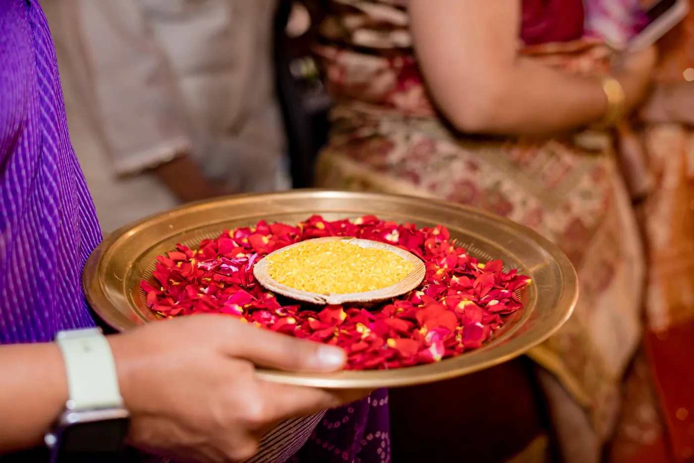 A brass plate of red petals and turmeric is held out by a hand