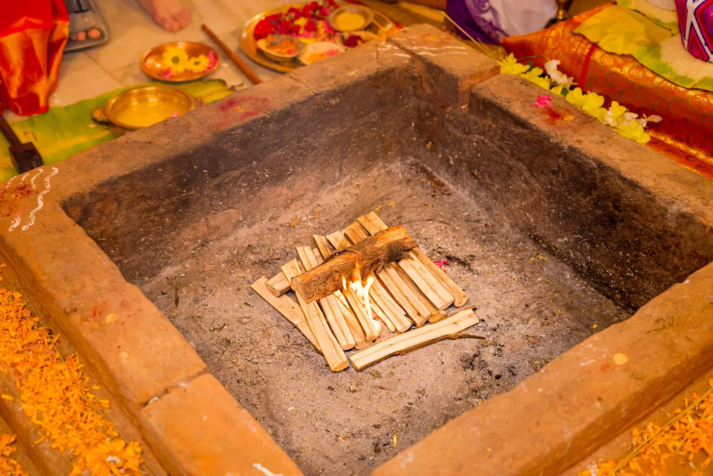 A square sacred fire pit holds stacked wooden sticks and marigolds