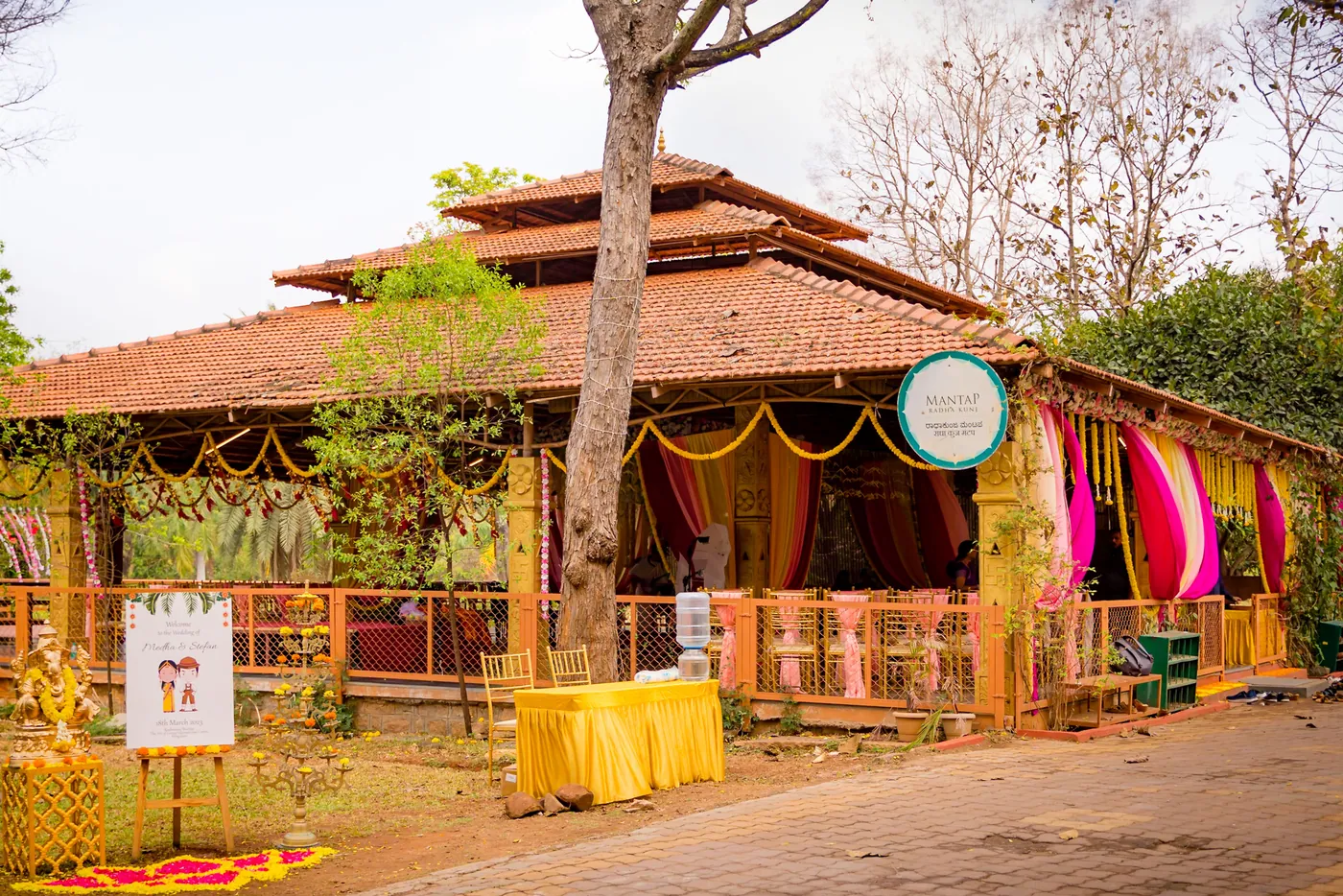 A wooden pavilion with a tiled roof and pink and yellow drapes stands among trees at the ashram