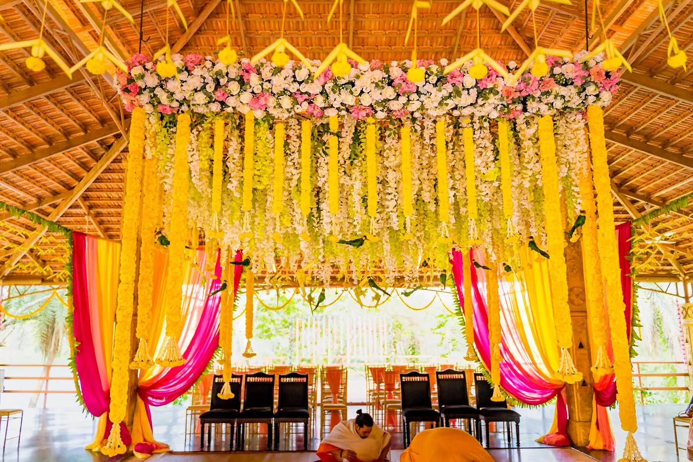 A mandap with hanging flower strings and yellow and pink drapes stands over rows of chairs