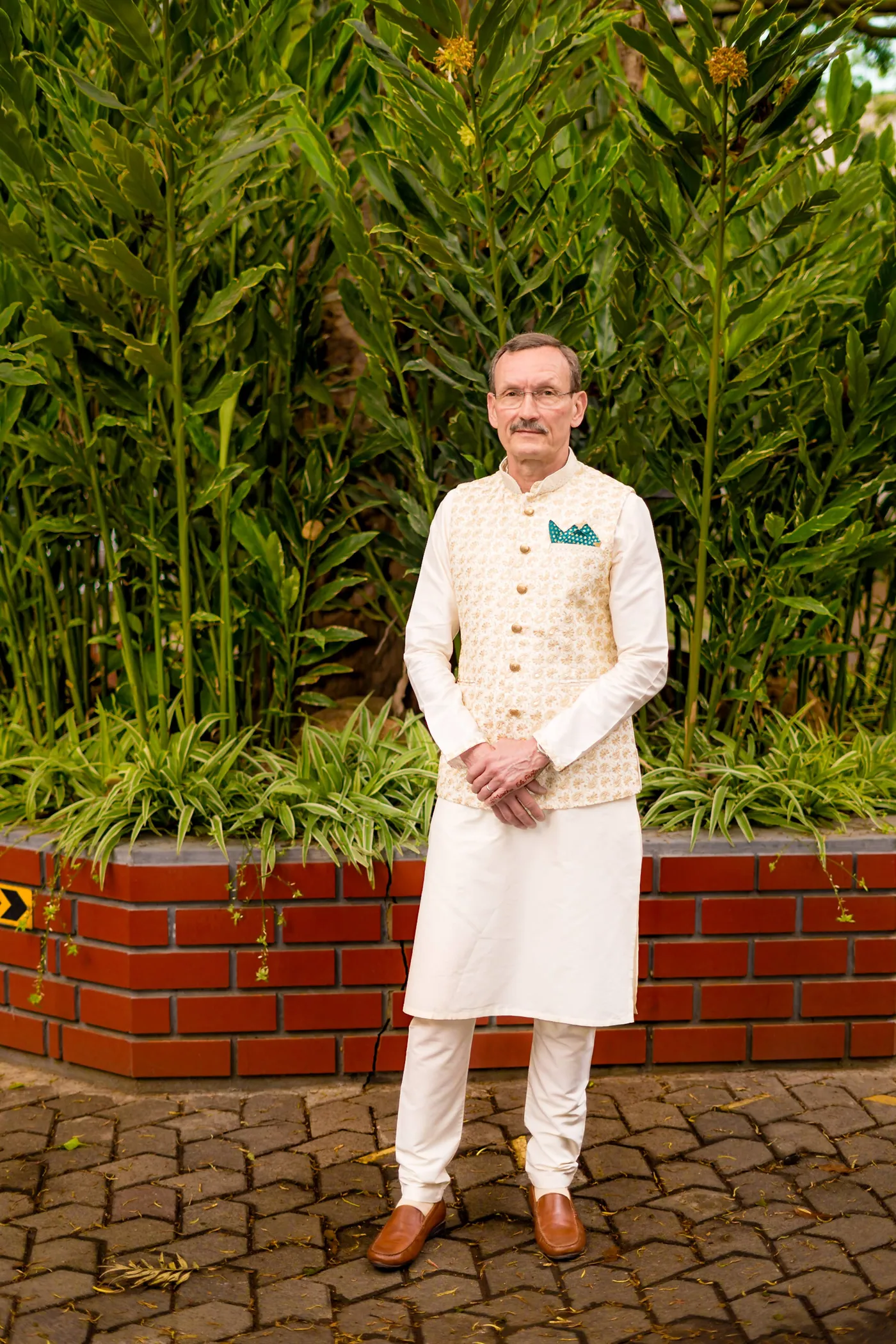 A man in a cream kurta and waistcoat stands in front of tall green plants and a brick wall