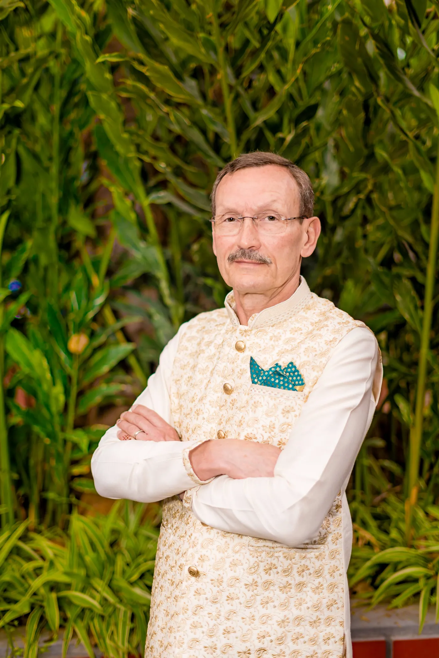 Stefan's father in a cream embroidered waistcoat, arms crossed, among green foliage