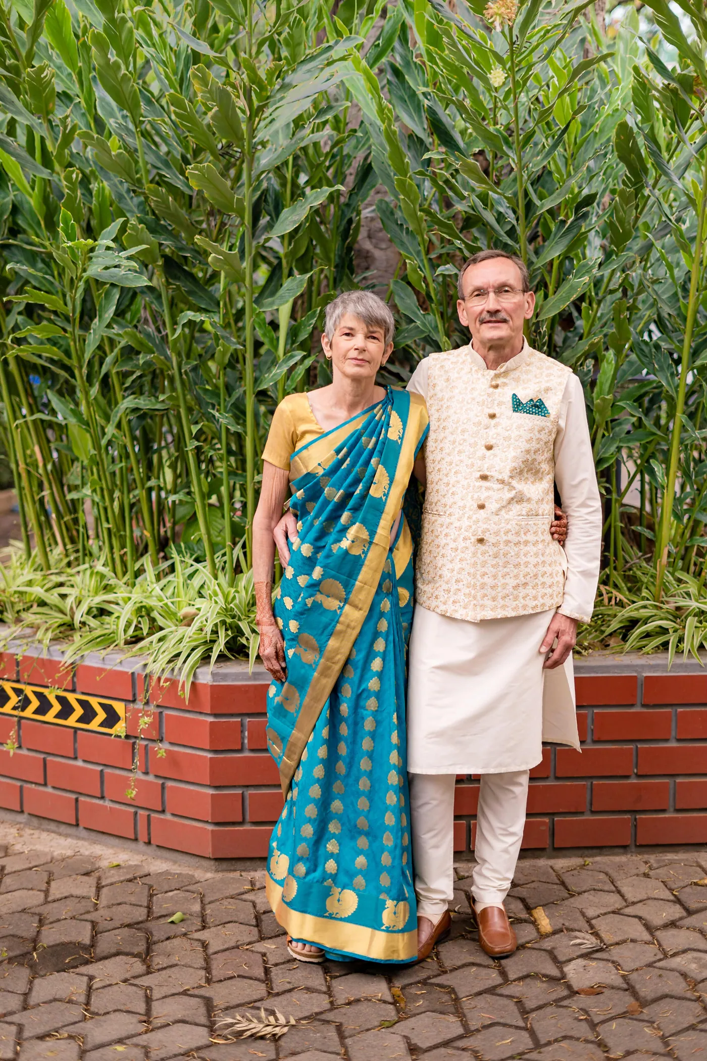 A woman in a teal sari and a man in a cream waistcoat pose together by a brick planter