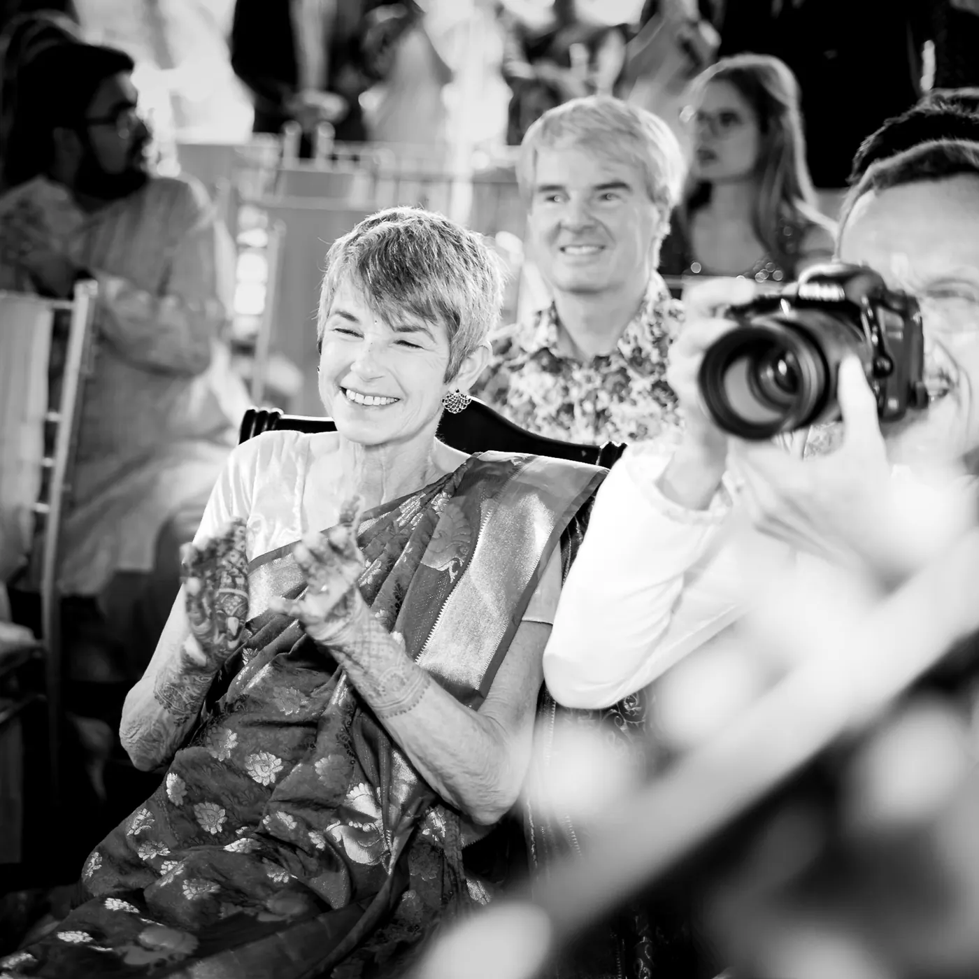 Black and white of a grey-haired woman smiling as a man photographs the ceremony