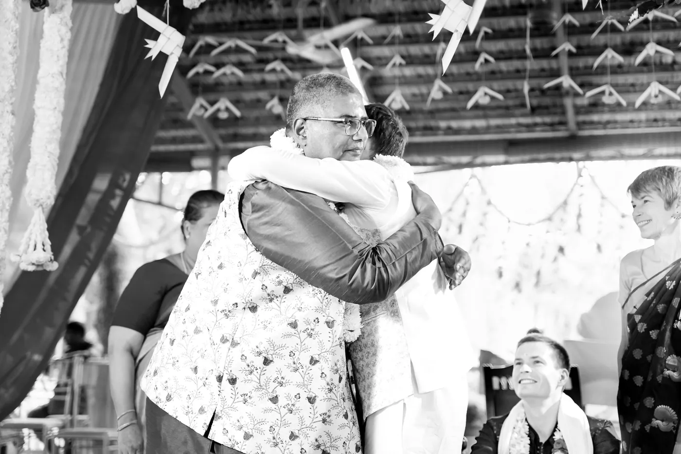 Black and white of two men embracing warmly under a thatched roof