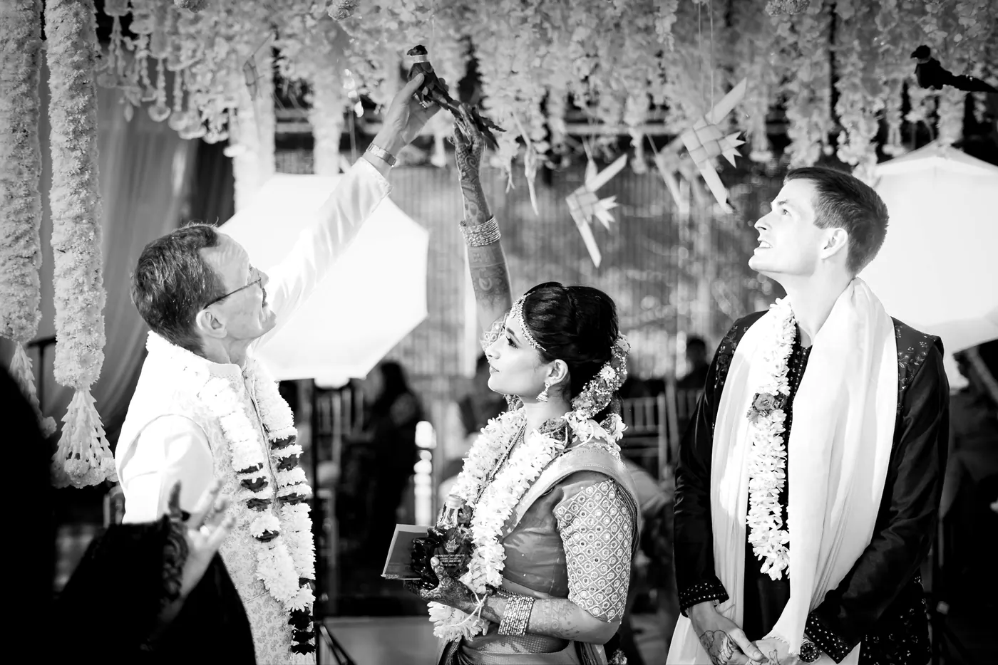 Black and white of a man in a white jacket speaking with Medha and Stefan in white garlands