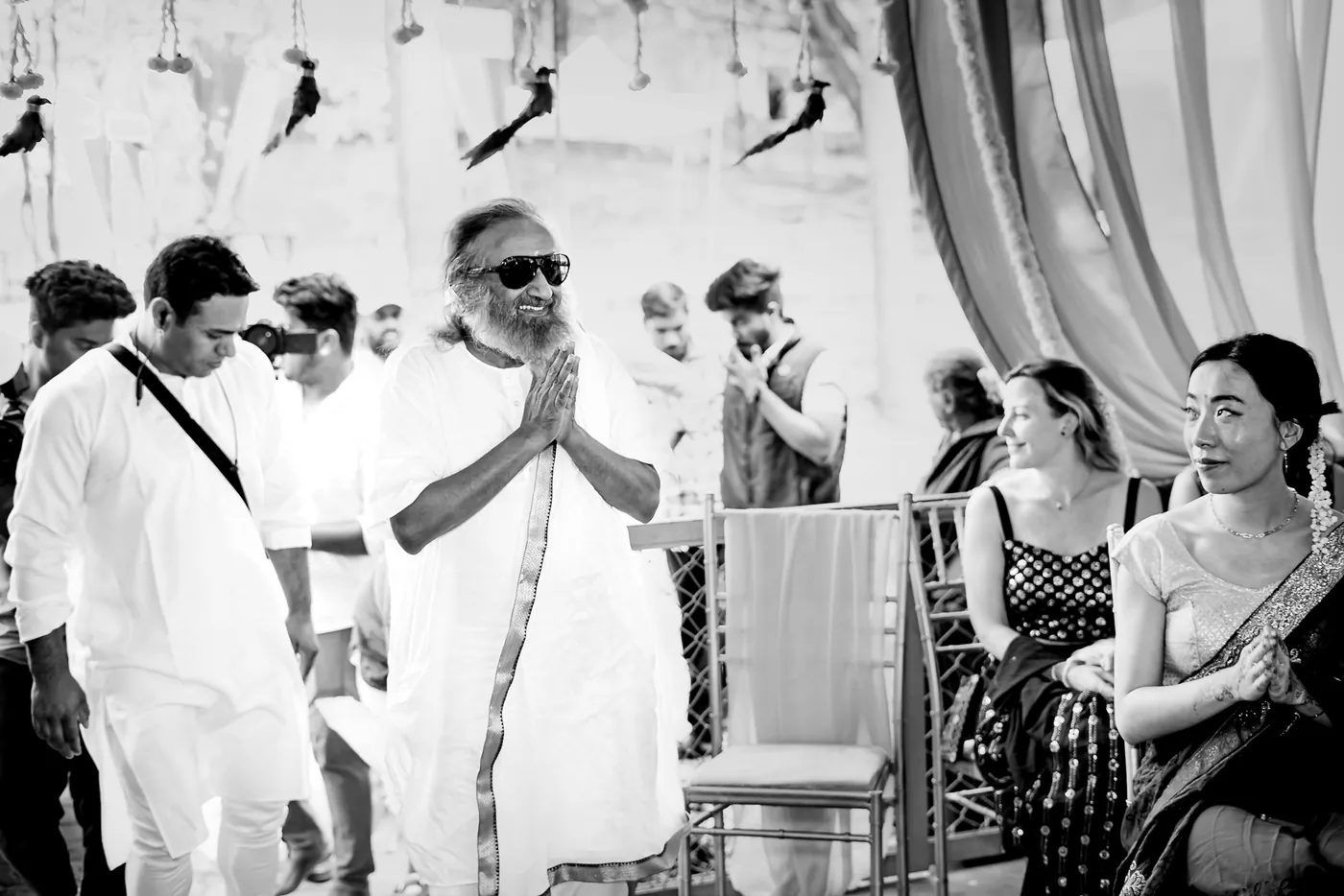 Black and white of a man in white with folded hands among guests
