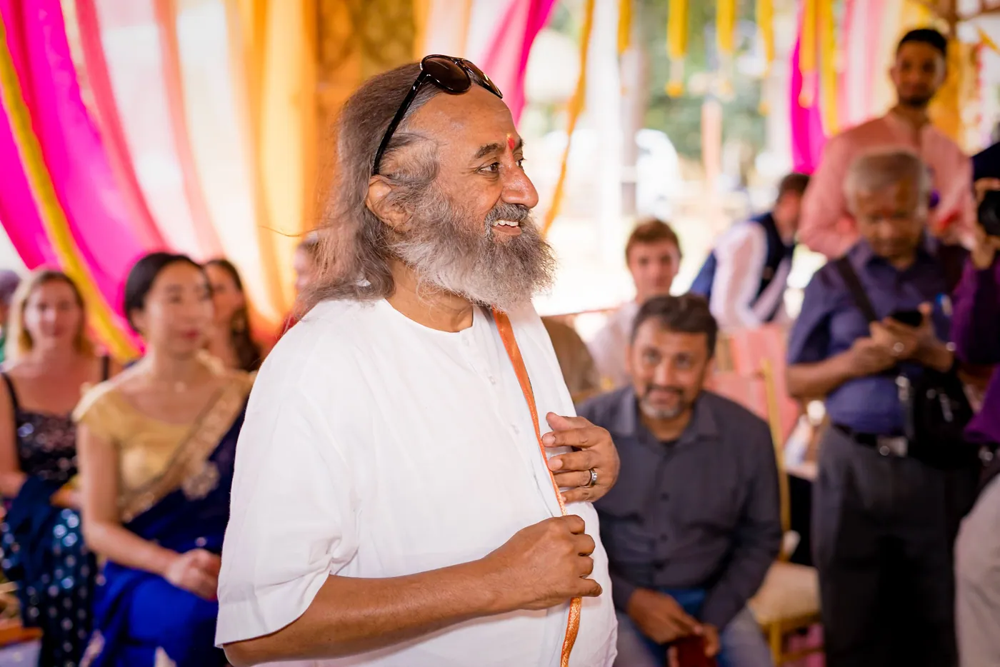A man in white robes with a long beard and lotus garland smiles among guests