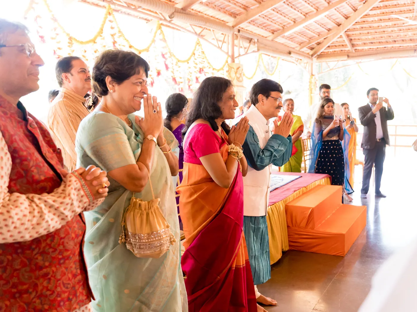 Guests stand with folded hands in a hall as a woman in green smiles toward the stage