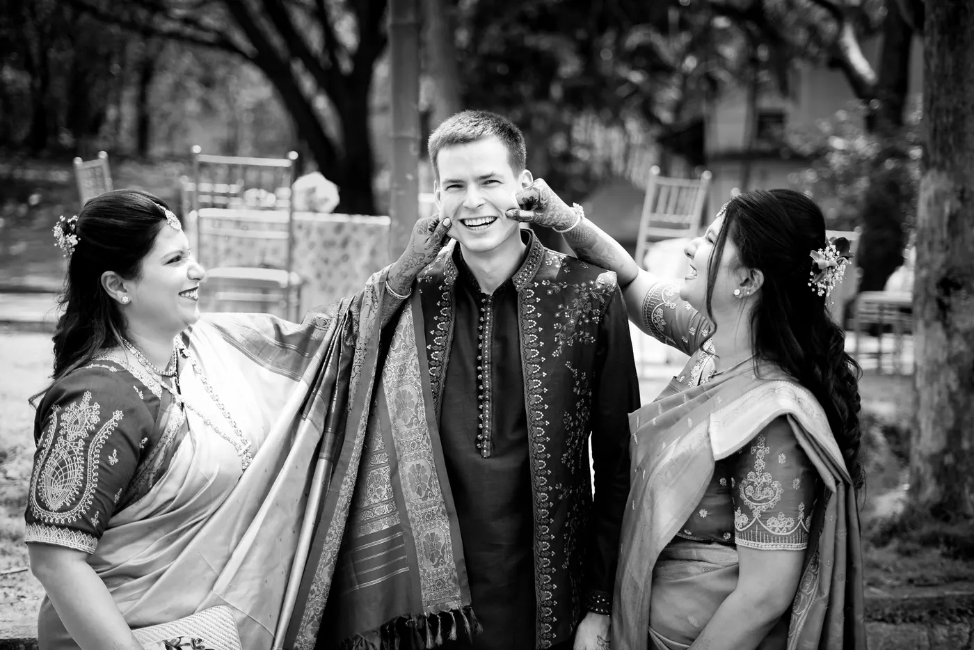 Two women playfully pinch Stefan's cheeks as he grins, with chairs in the garden behind