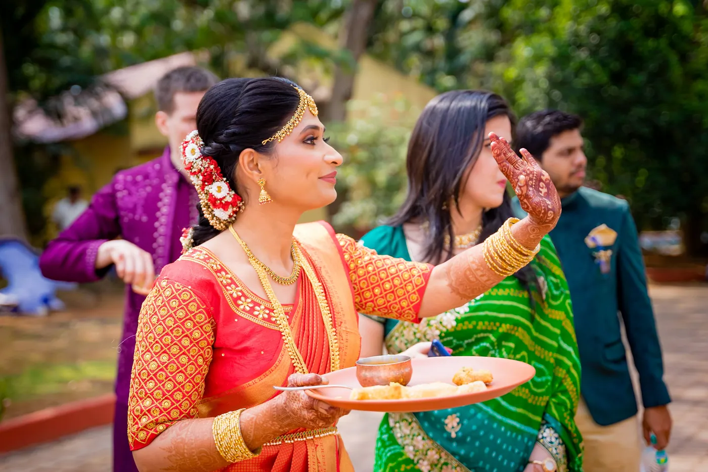 Medha in an orange blouse holds a plate of offerings while guests in bright clothes stand behind her