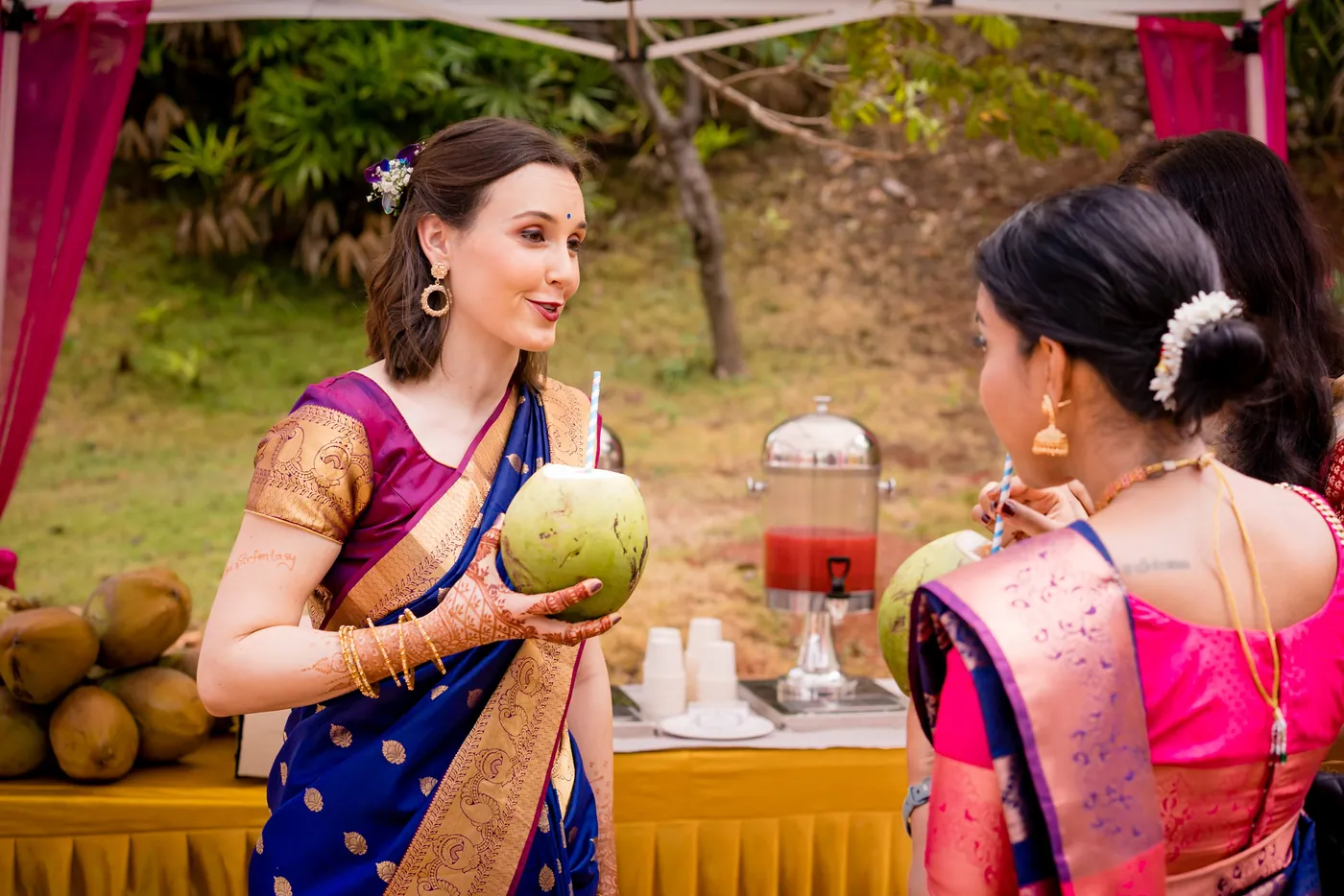 A woman holds a coconut and a drink while chatting with a woman in a green blouse