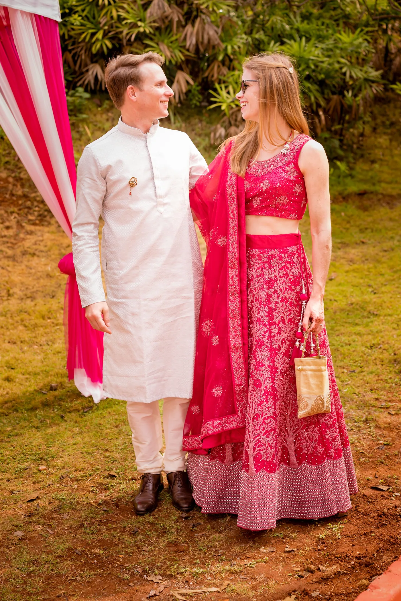 A man in a white kurta and a woman in a pink lehenga pose together under trees