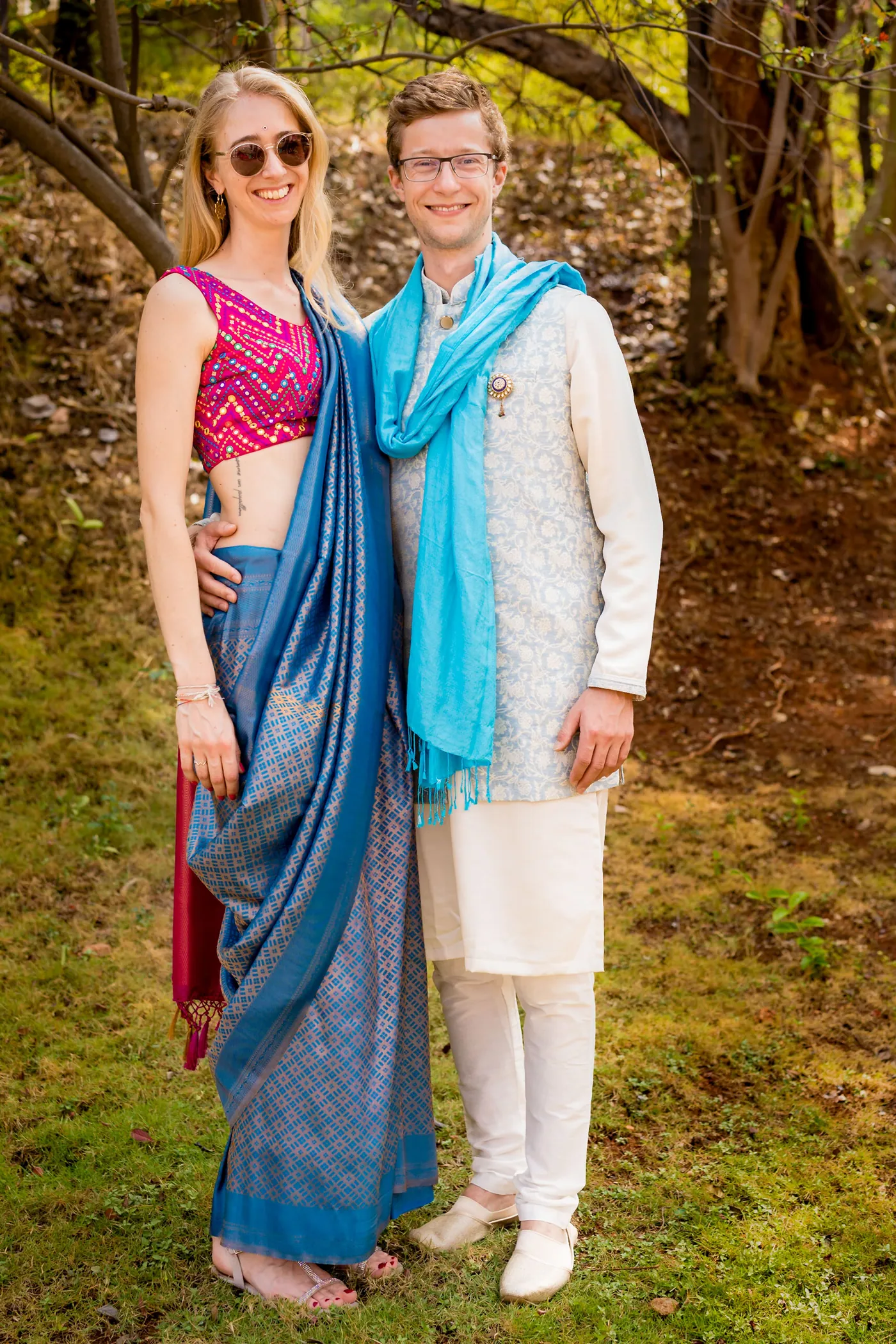 A woman in a blue stole and a man in a cream kurta smile under a tree