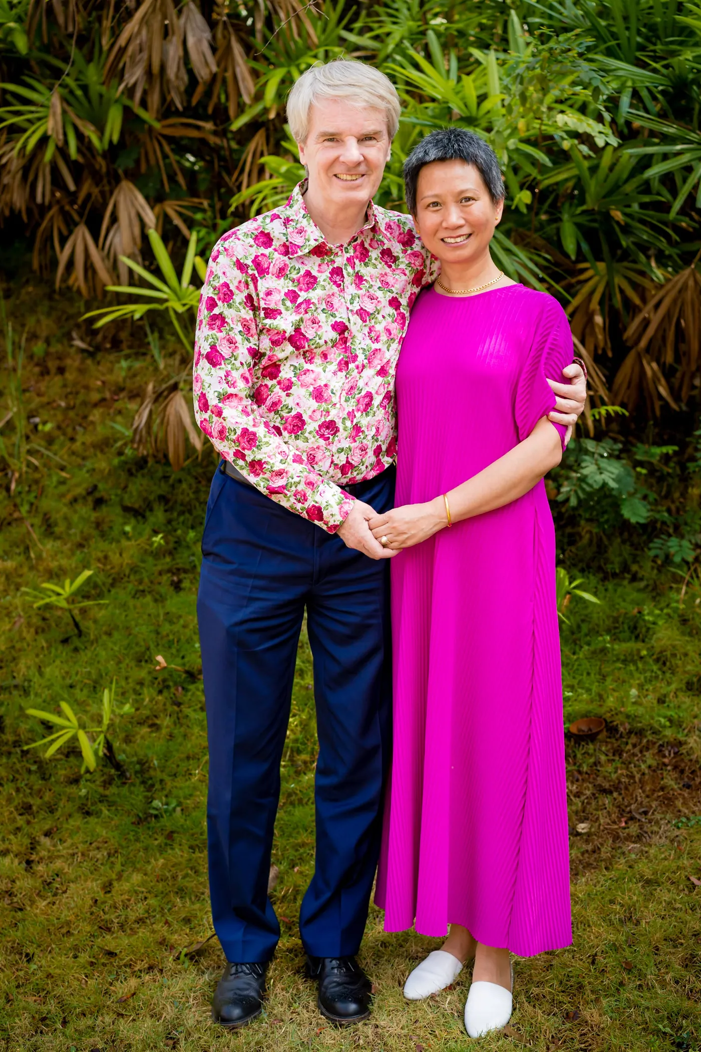 A man in a floral shirt and a woman in a pink dress hold each other in a garden