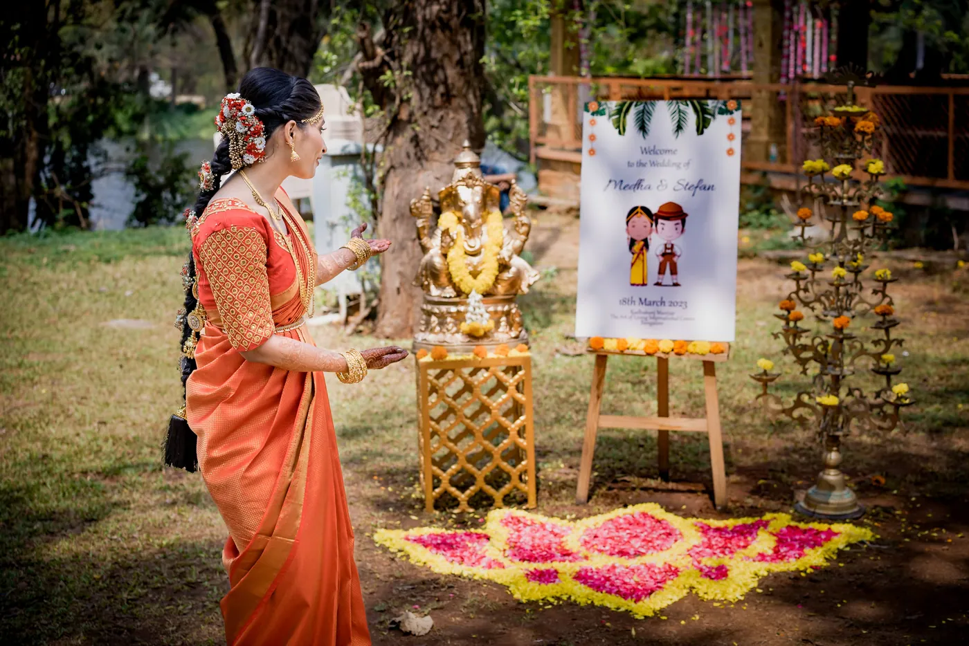 A woman in an orange sari places an offering near a Ganesha statue beside a welcome board