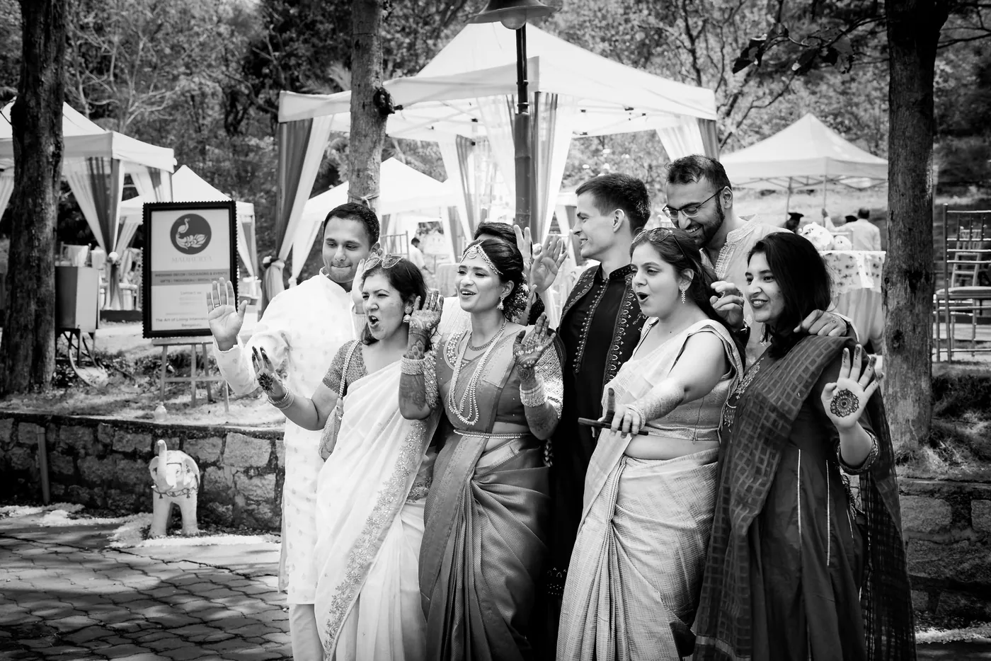 Black and white of guests in saris and suits gathered and smiling near white tents