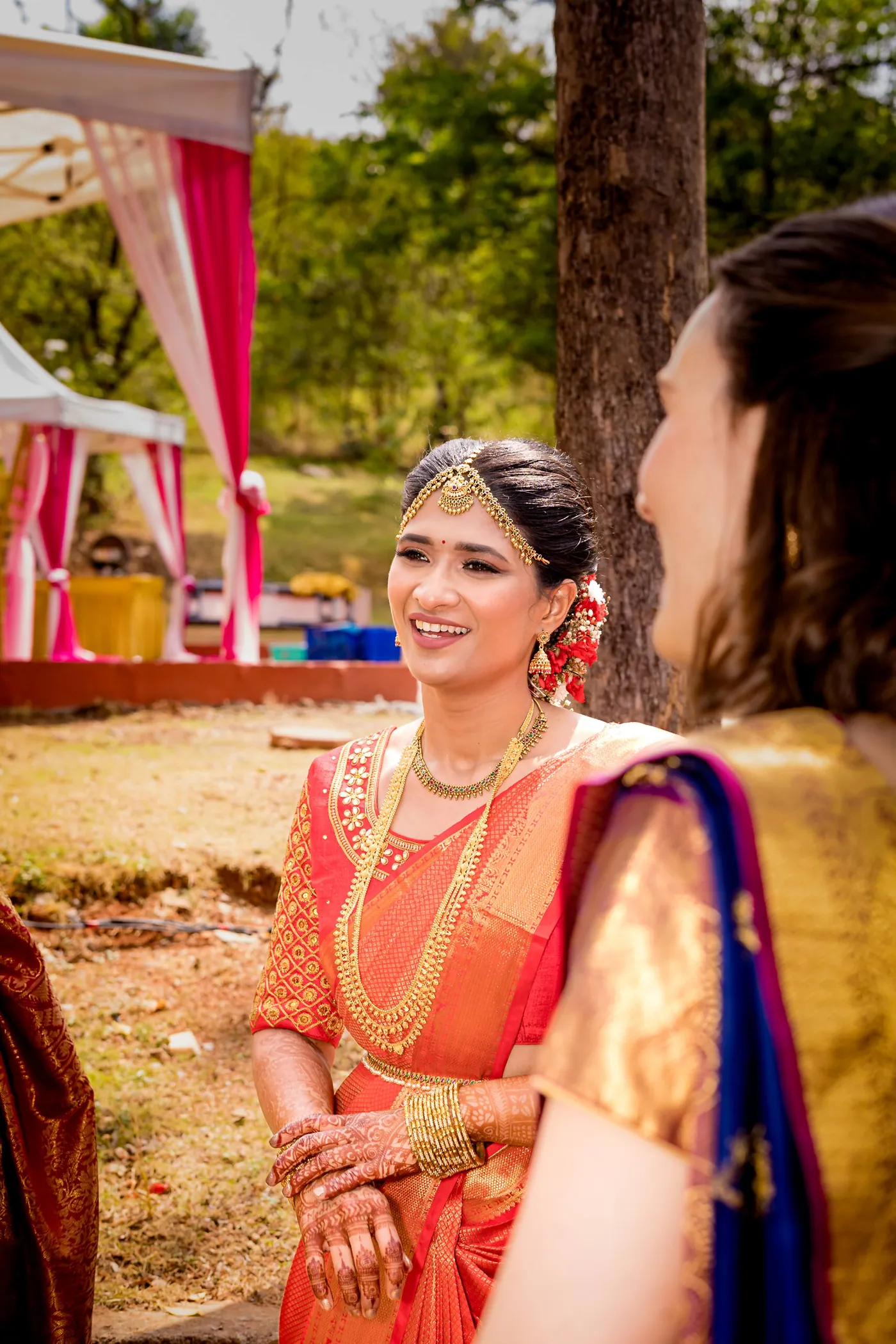 Medha in an orange sari and gold jewellery smiles while talking to a guest