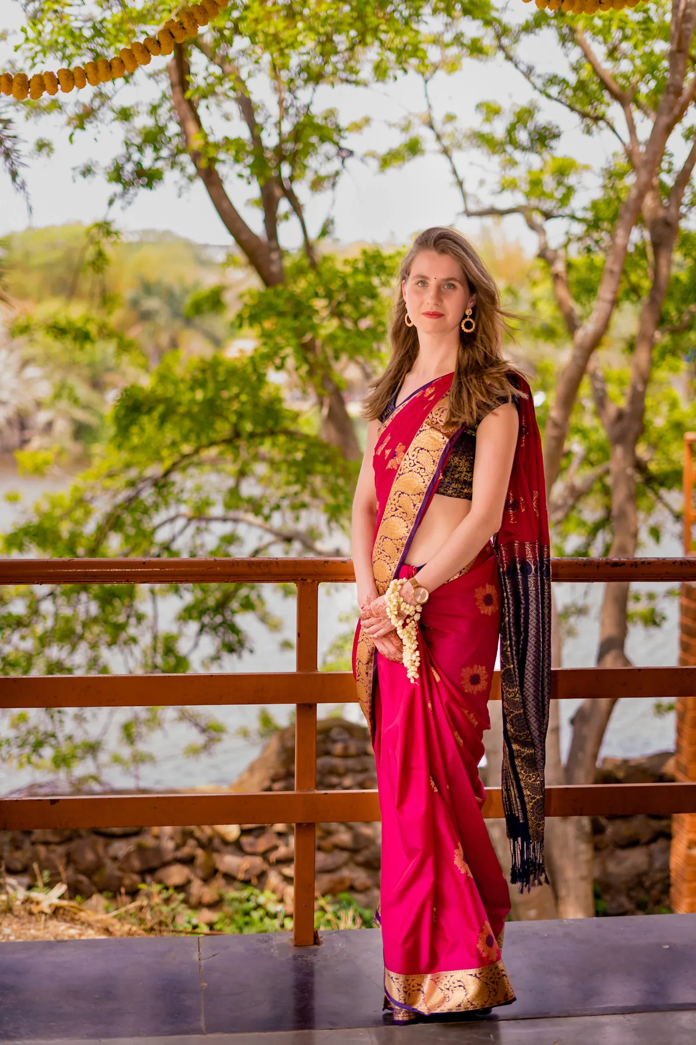A woman in a red sari stands at a wooden railing with trees and the lake behind her