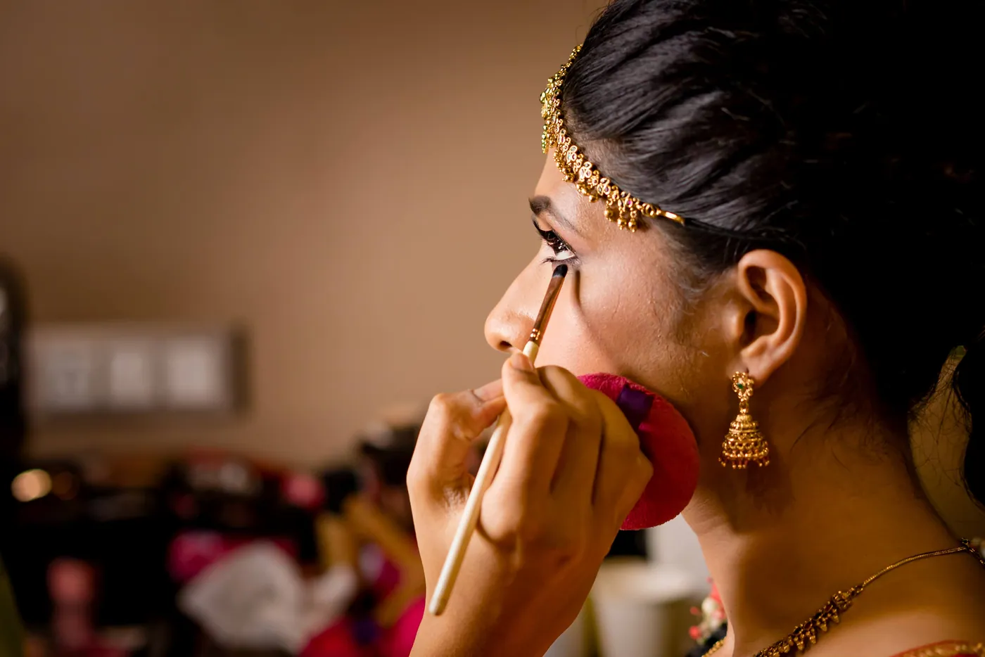 A makeup artist applies kohl to Medha's eye as she holds a small mirror in a warm room