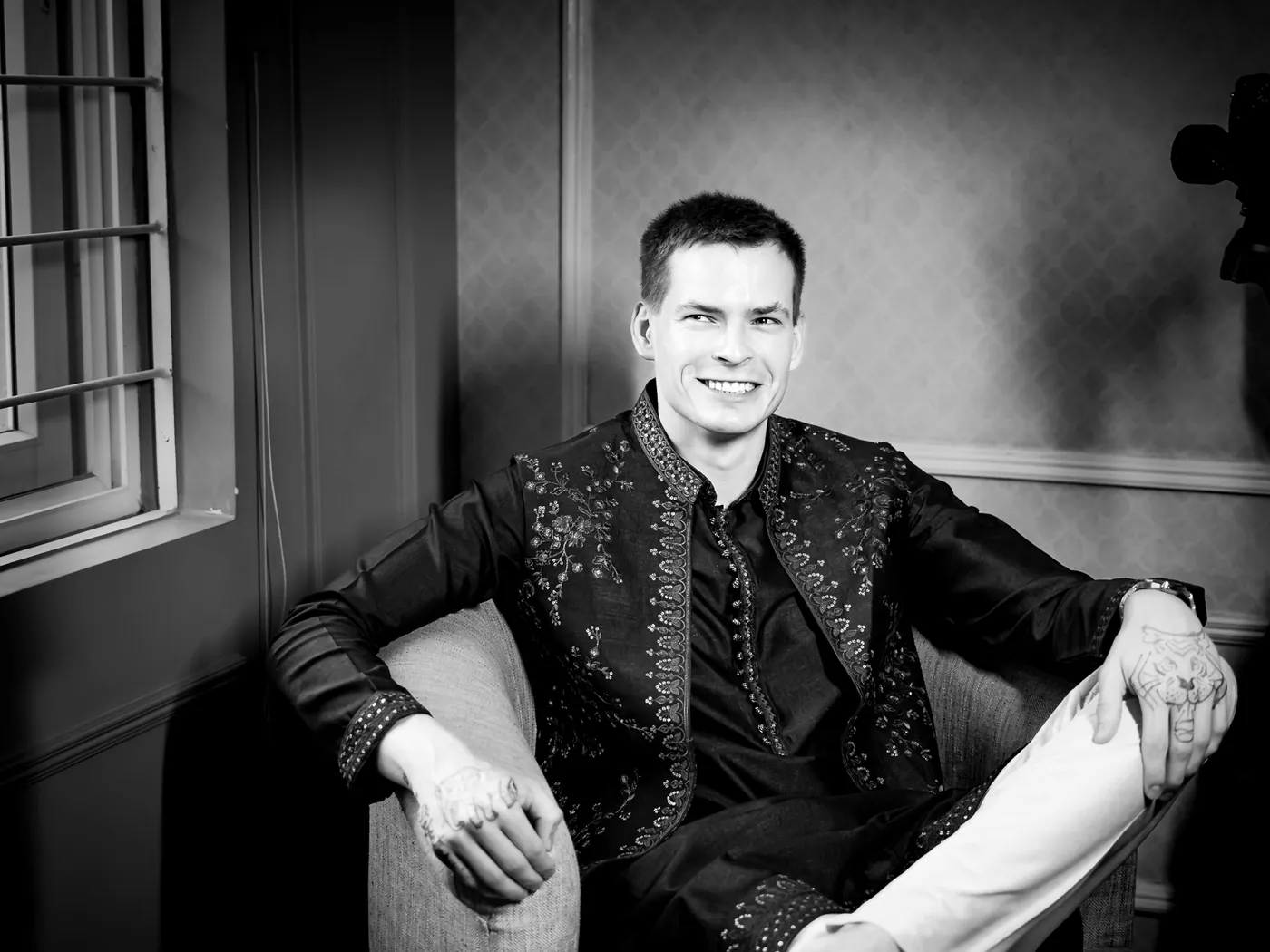 Black and white of Stefan relaxed in an armchair in an embroidered jacket, smiling at the camera