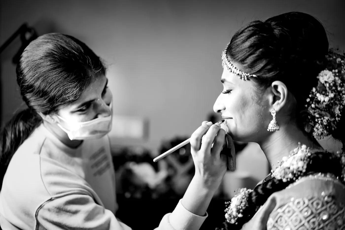 Black and white of a masked makeup artist applying lipstick to Medha, who wears a headpiece and earrings