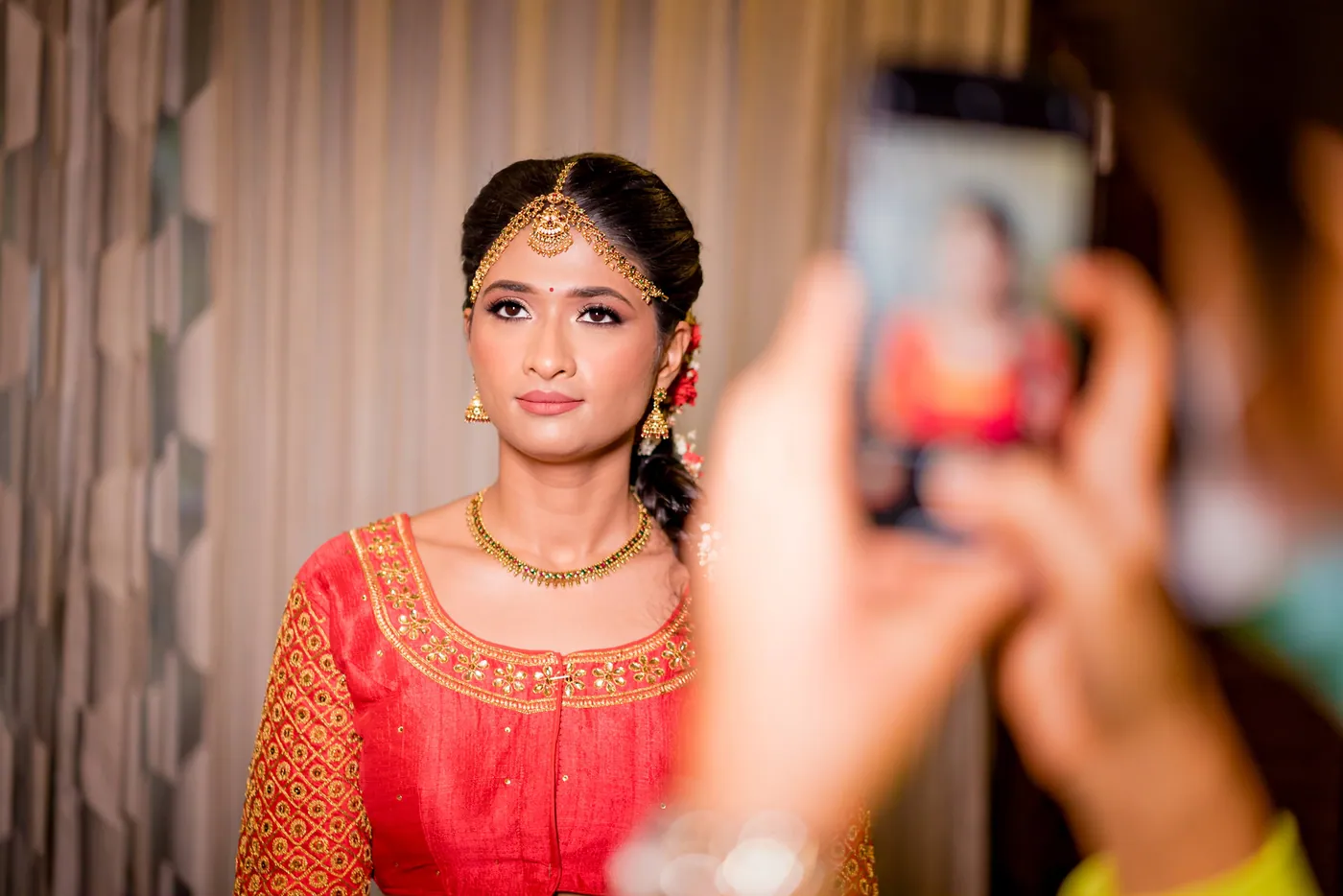 Medha in an orange blouse and gold headpiece looks calmly ahead while a guest photographs her with a phone